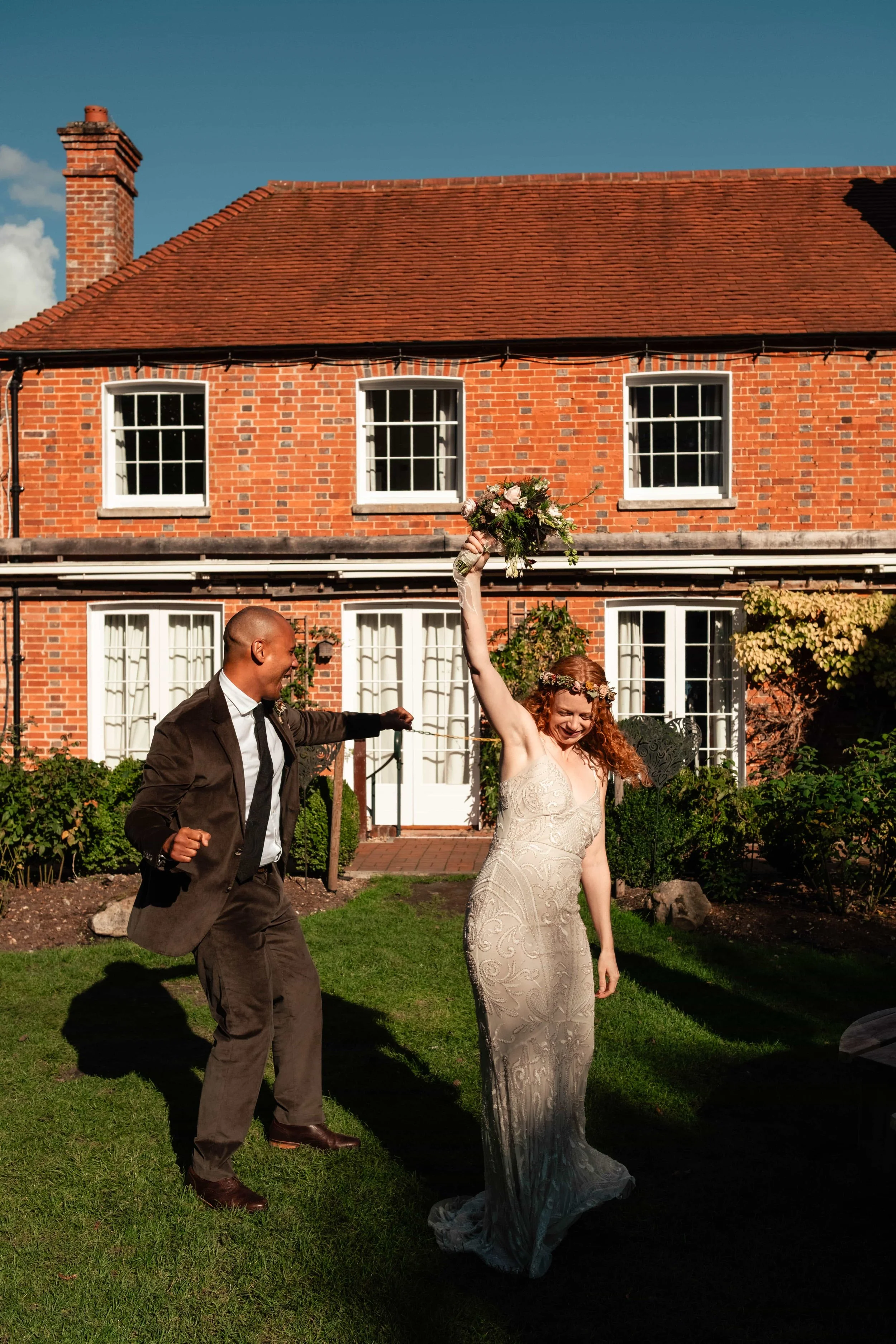 A bride in a white wedding dress with a floral headband celebrating outside a red brick house with a man in a brown suit holding a bouquet, sunlight casting shadows on the grass.