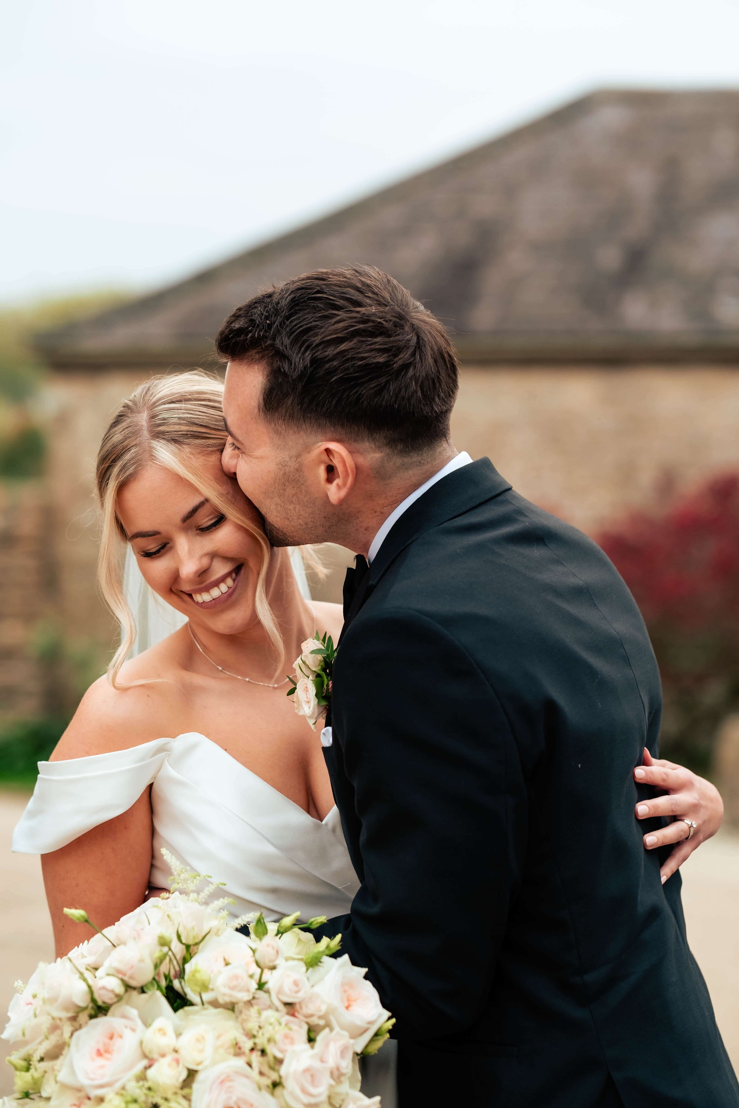 Bride and groom sharing a joyful moment, with the bride holding a bouquet of pink and white flowers, outdoors on a cloudy day.
