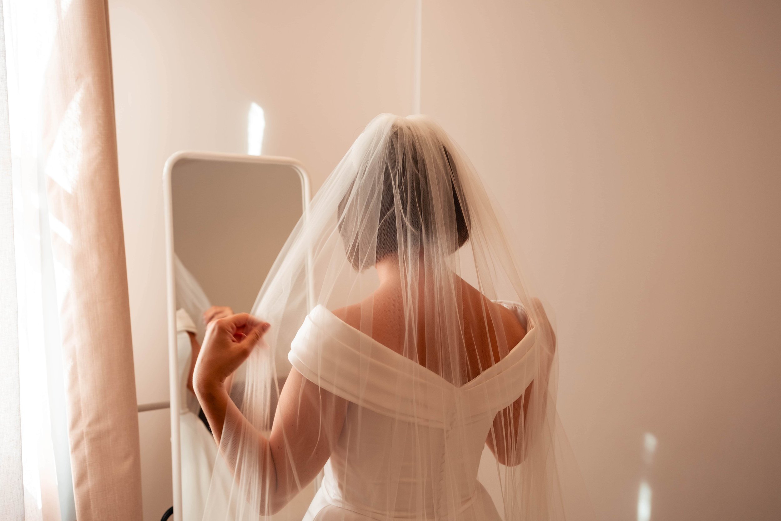 A bride in a white dress and veil looking into a mirror.