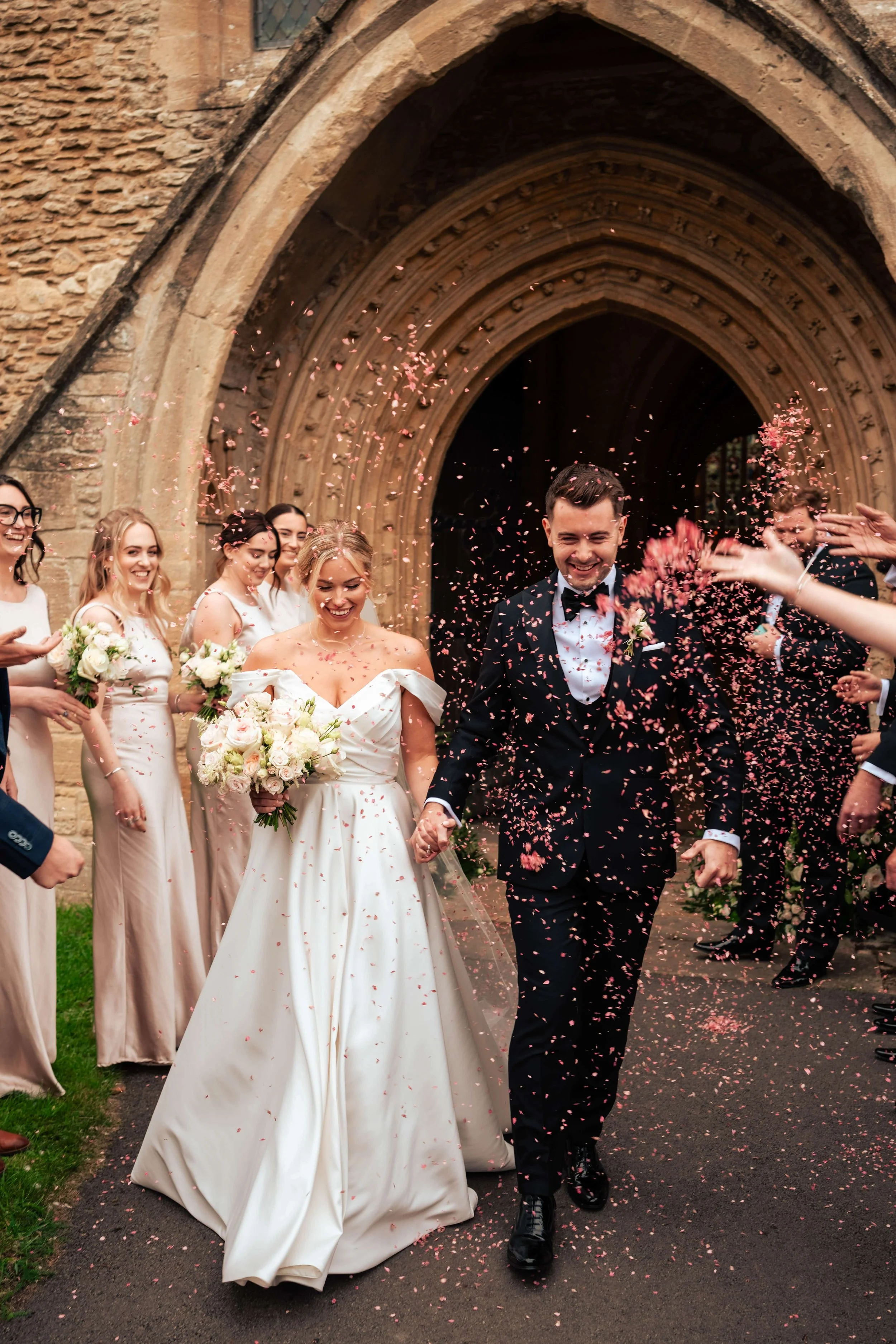 A bride and groom walking hand in hand outside a stone church, surrounded by friends and family throwing pink confetti during their wedding celebration.