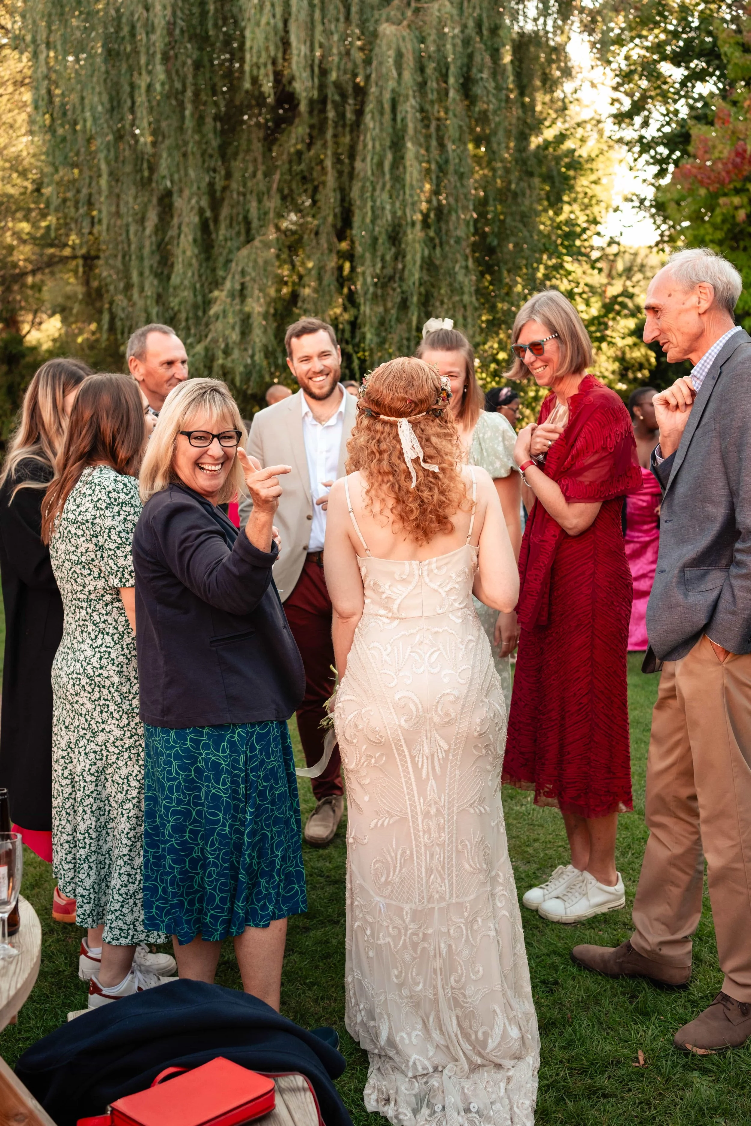 People gathered outdoors at sunset, smiling and talking, with a woman in a light-colored wedding dress at the center, surrounded by friends and family.