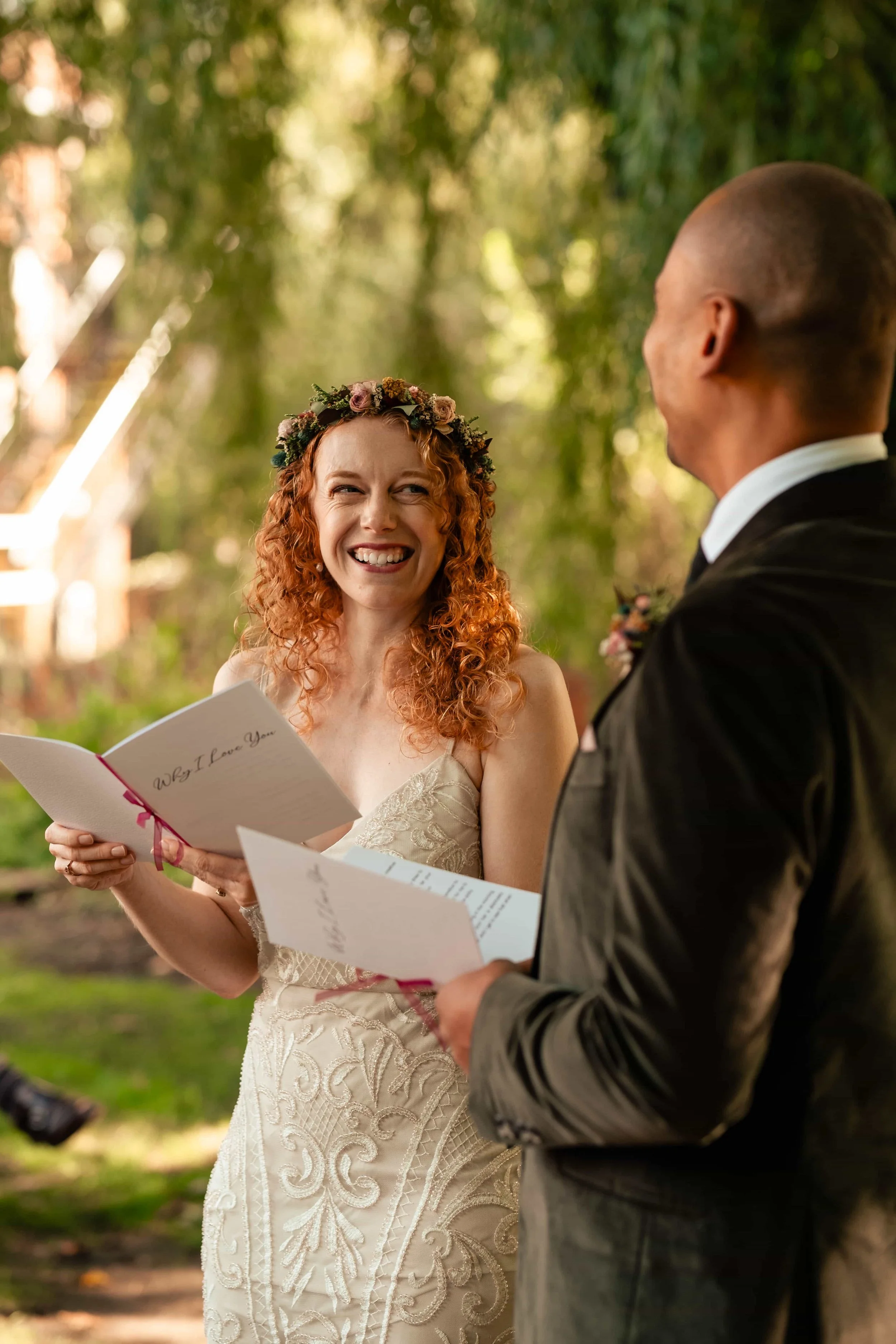 A woman with curly red hair wearing a wedding dress and a floral crown smiles at a man in a suit during a wedding ceremony outdoors.