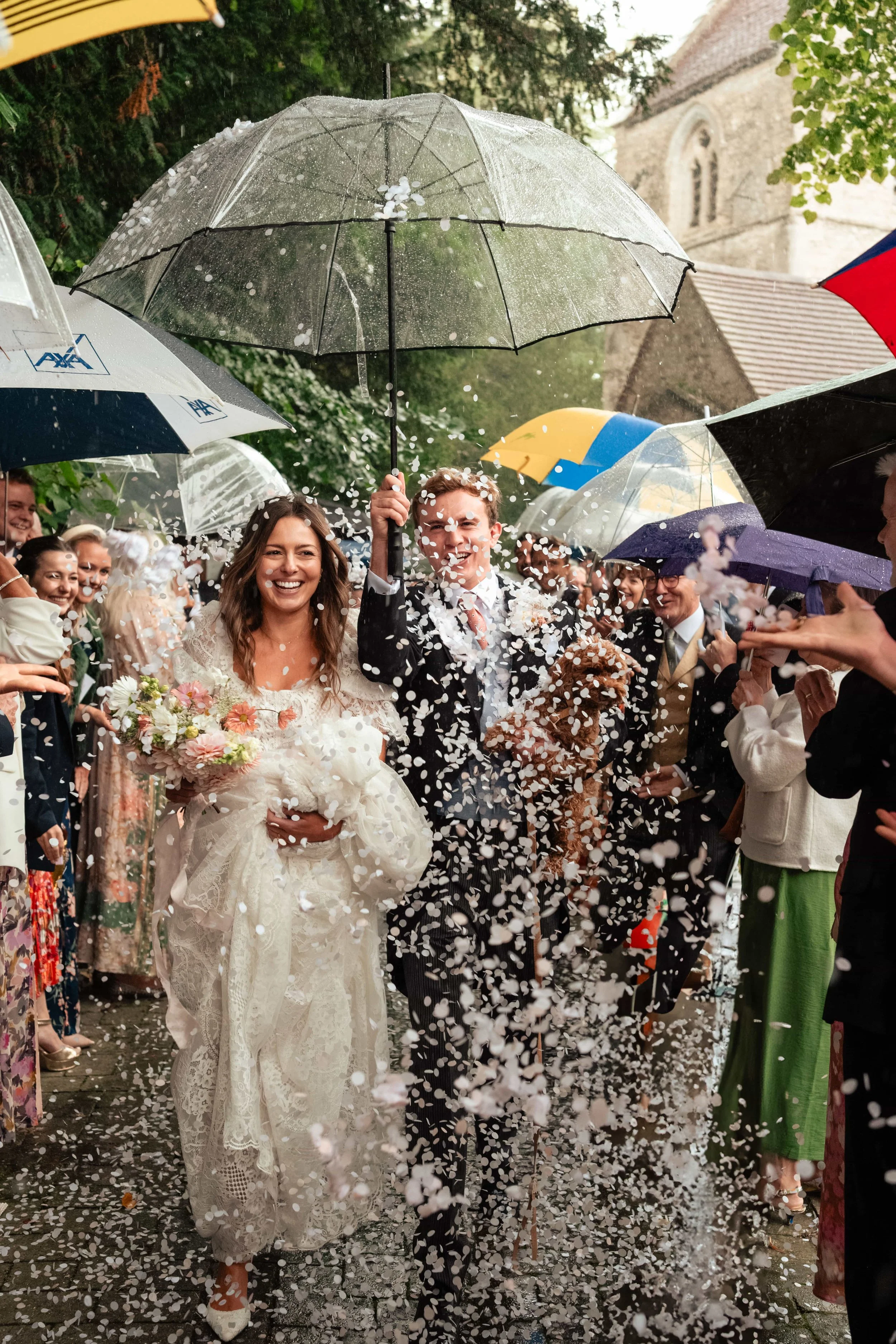 Bride and groom walking down a confetti tunnel, holding an umbrella and their dog, smiling on their wedding day.