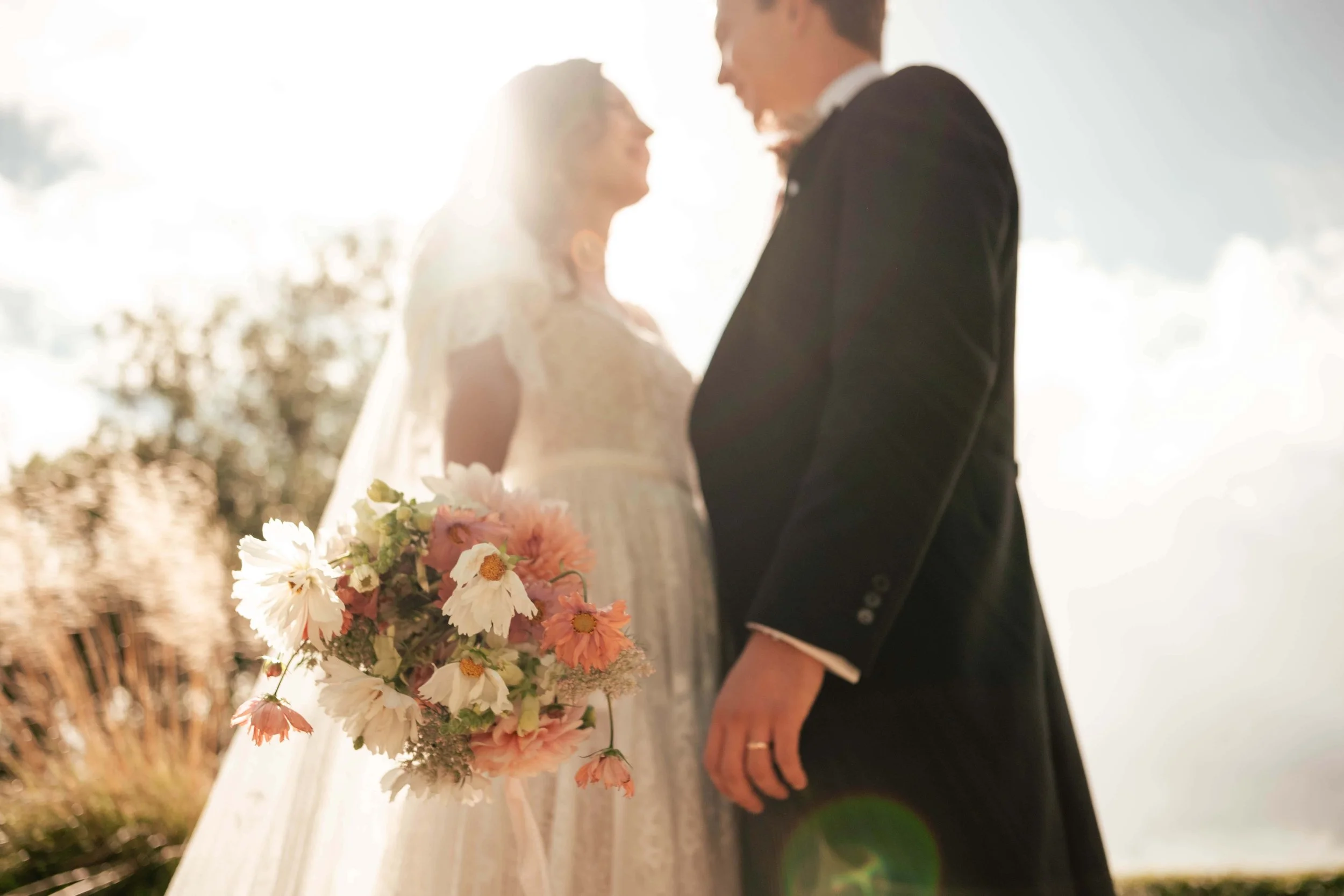 A bride and groom standing outdoors during sunset, with the bride holding a bouquet of pink and white flowers.