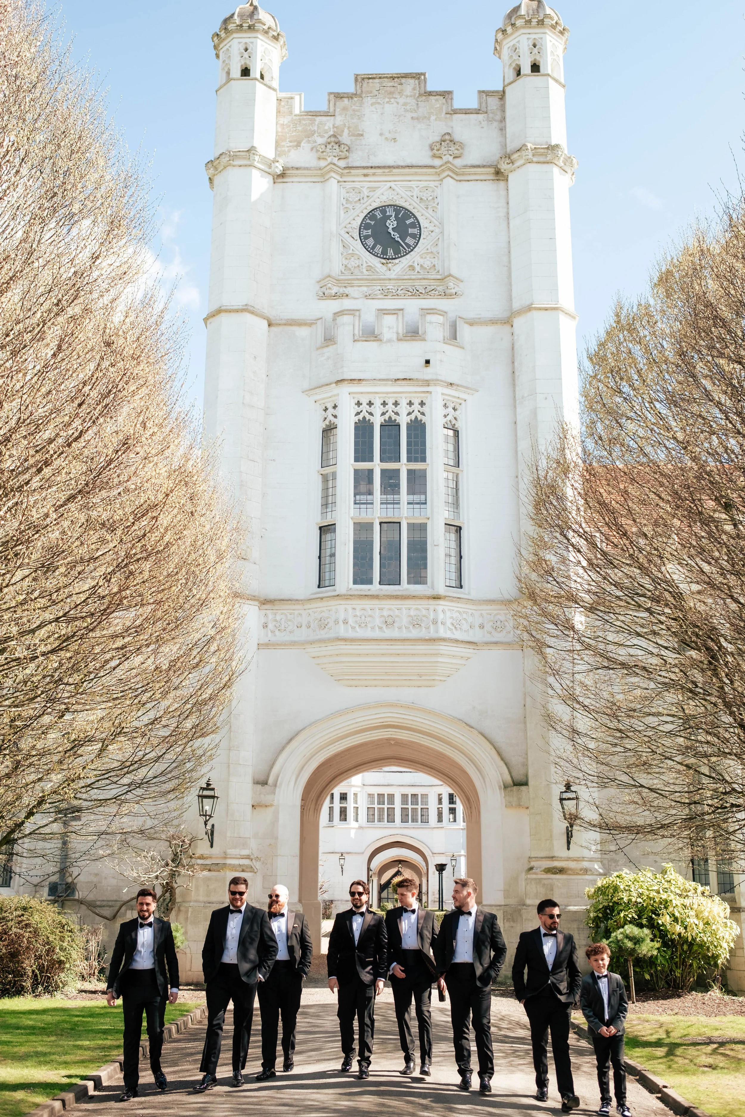 A groom and his groomsmen dressed in tuxedos and bow ties walking in front of a historic white building with a clock tower, surrounded by trees and bushes on a sunny day.