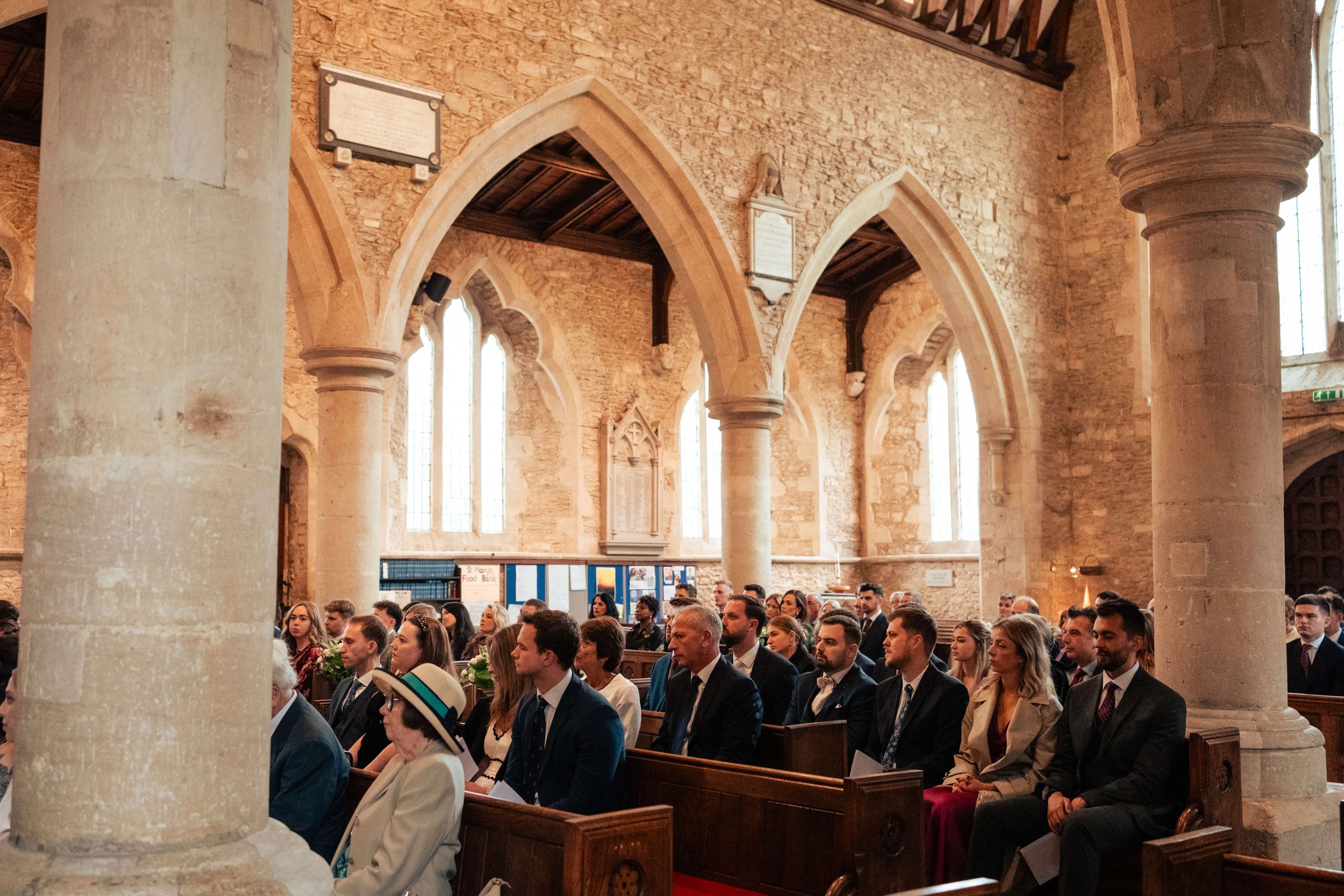People sitting in pews inside a stone church with high vaulted ceilings and tall stained glass windows.