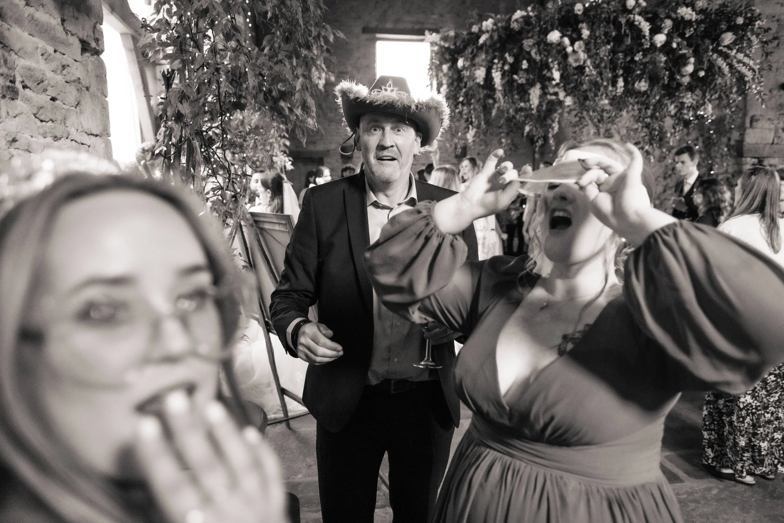 A person at a lively party wearing a cowboy hat looks surprised as a woman in a dress plays with a party blowout, with a crowd of guests and decorated walls in the background.