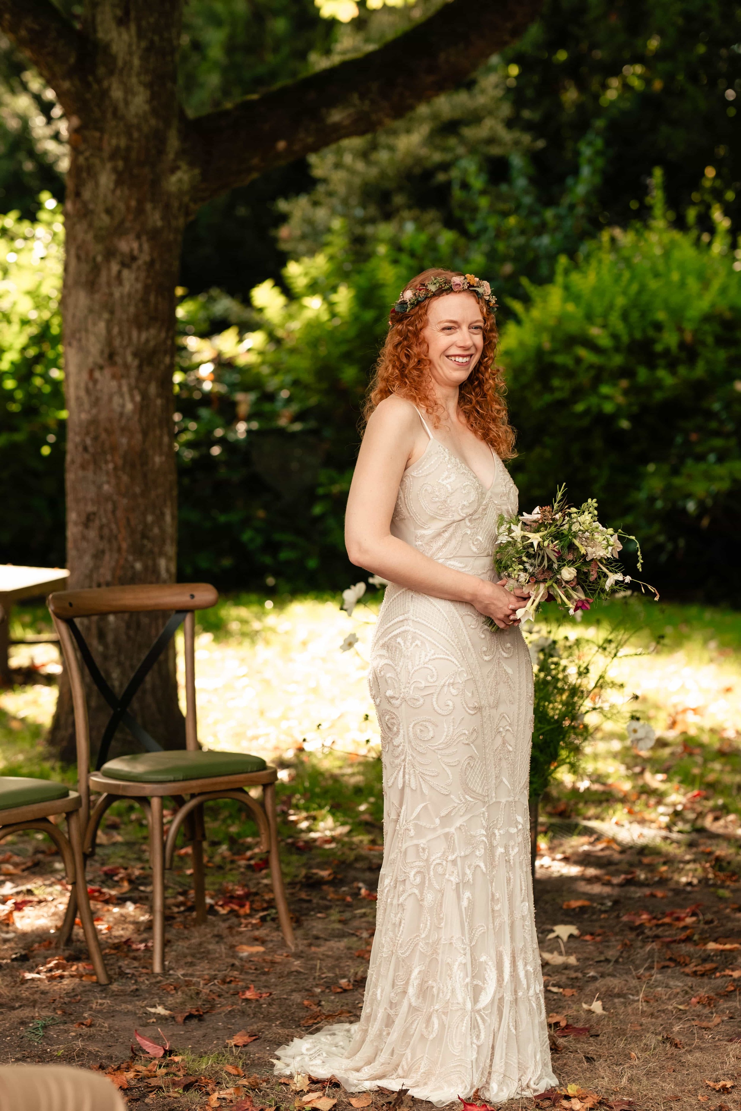 A woman in a romantic off-white lace wedding dress with spaghetti straps, standing outdoors under a large tree, holding a bouquet of flowers, smiling happily.