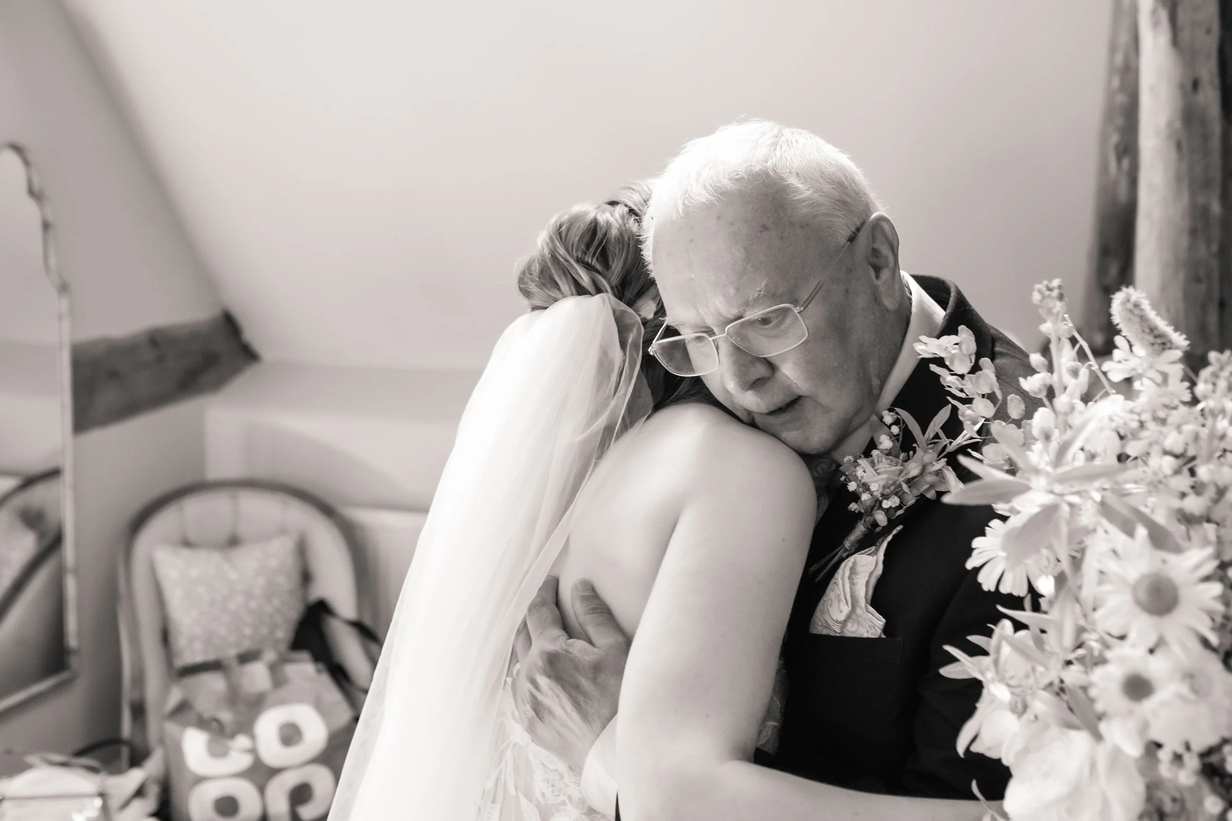 The father of the bride hugging his daughter on her wedding day, in a heartfelt embrace with flowers in the foreground.