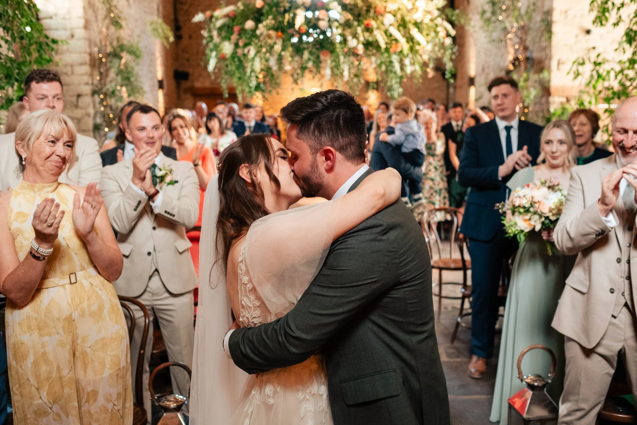 A bride and groom kiss in the center of a wedding reception with guests clapping and watching, decorated with hanging flowers and warm lighting.
