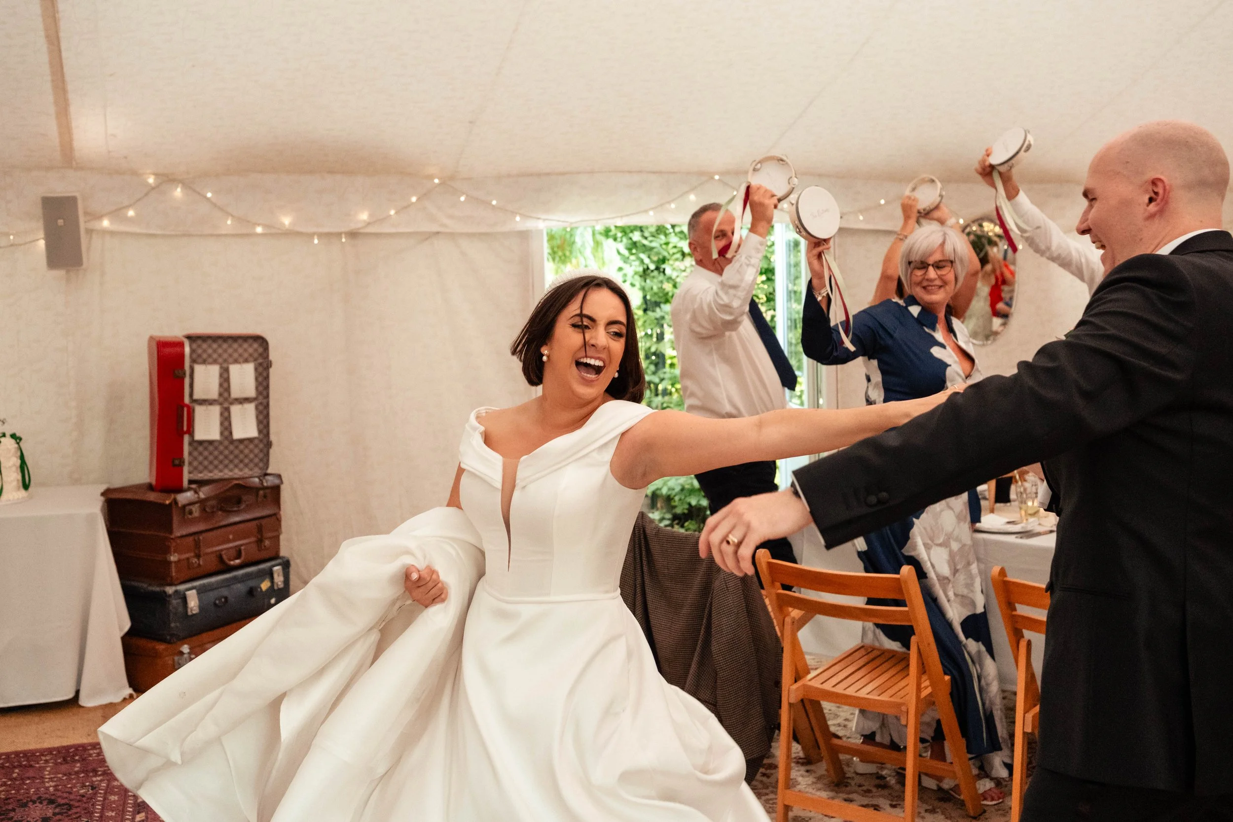 A bride and groom dancing at their wedding reception, with guests playing tambourines in the background.