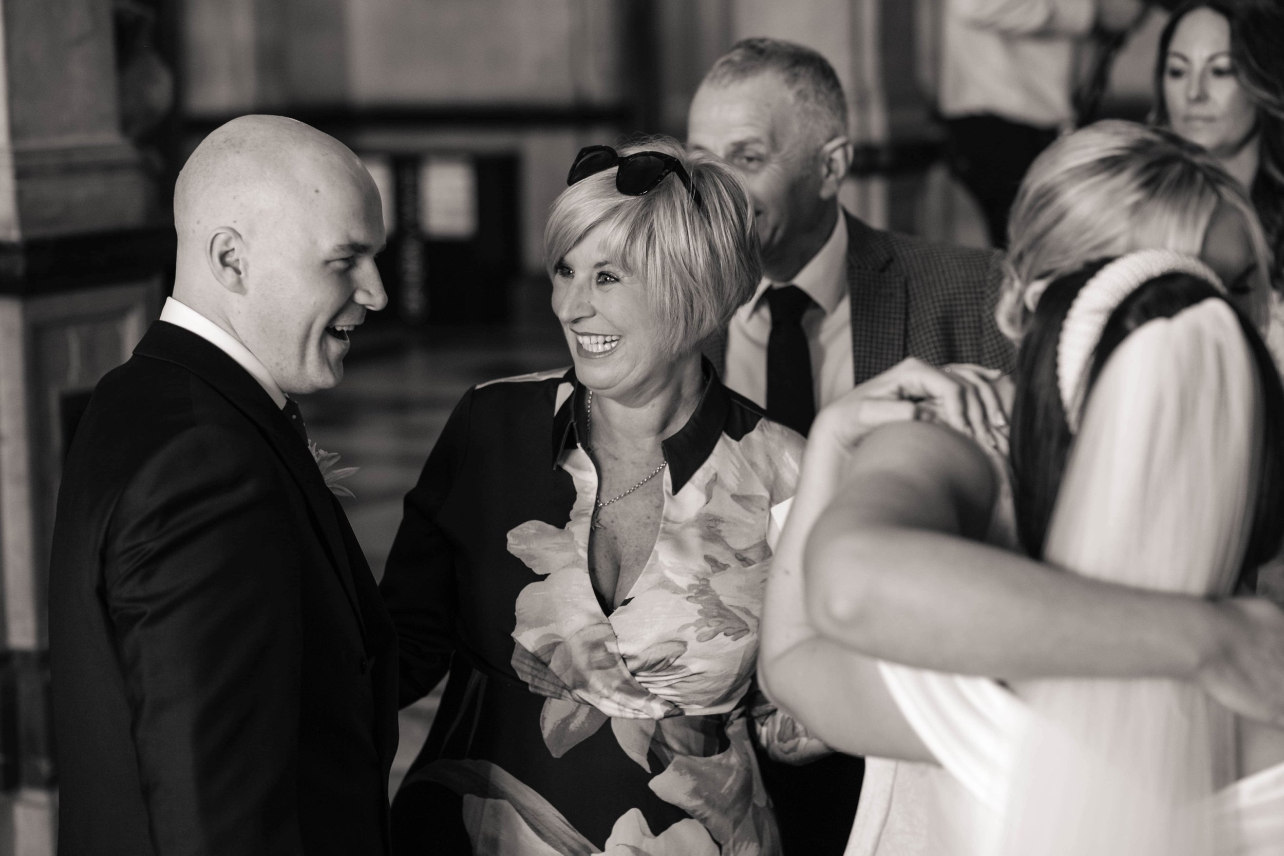 A group of people at a wedding in Oxford Town Hall, with a man in a suit and a woman with sunglasses on her head smiling and talking to each other.