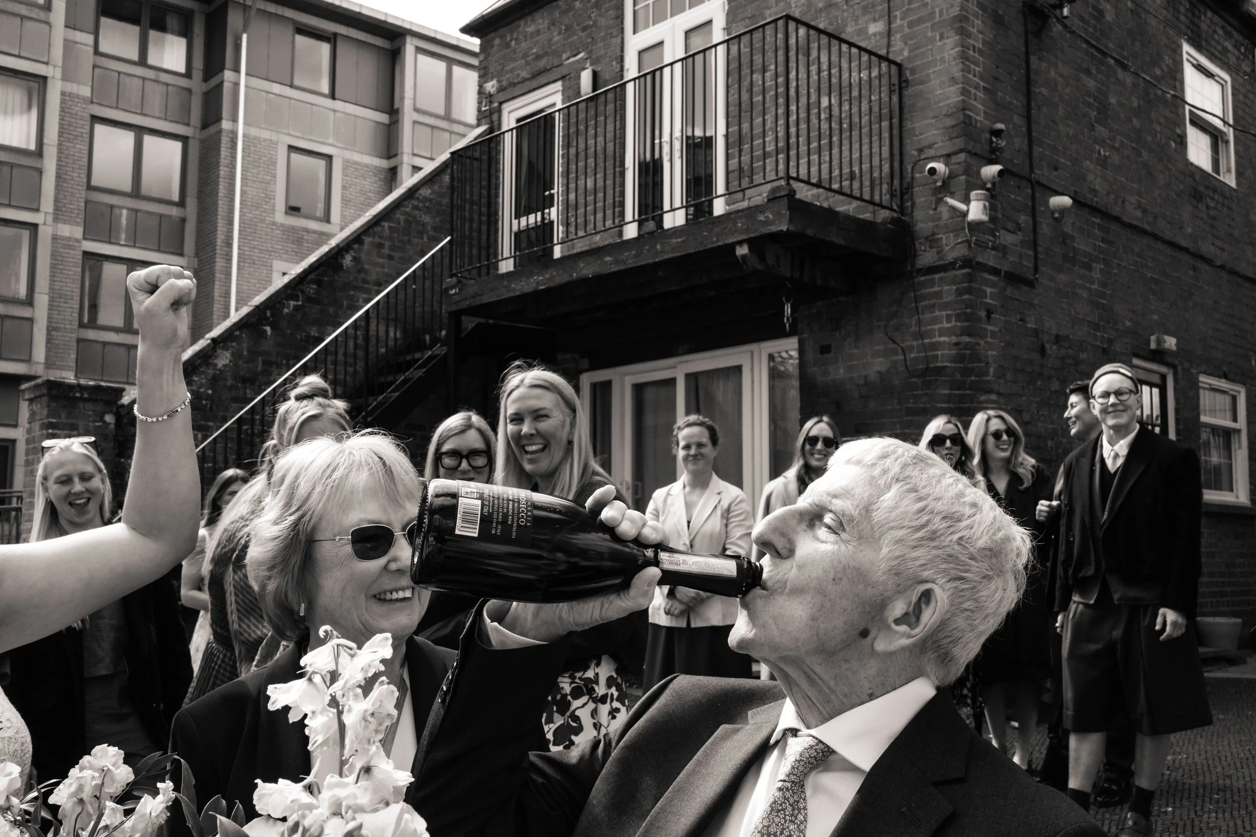 Grandpa drinking from a bottle of Prosecco surrounded by laughing wedding guests outdoors.