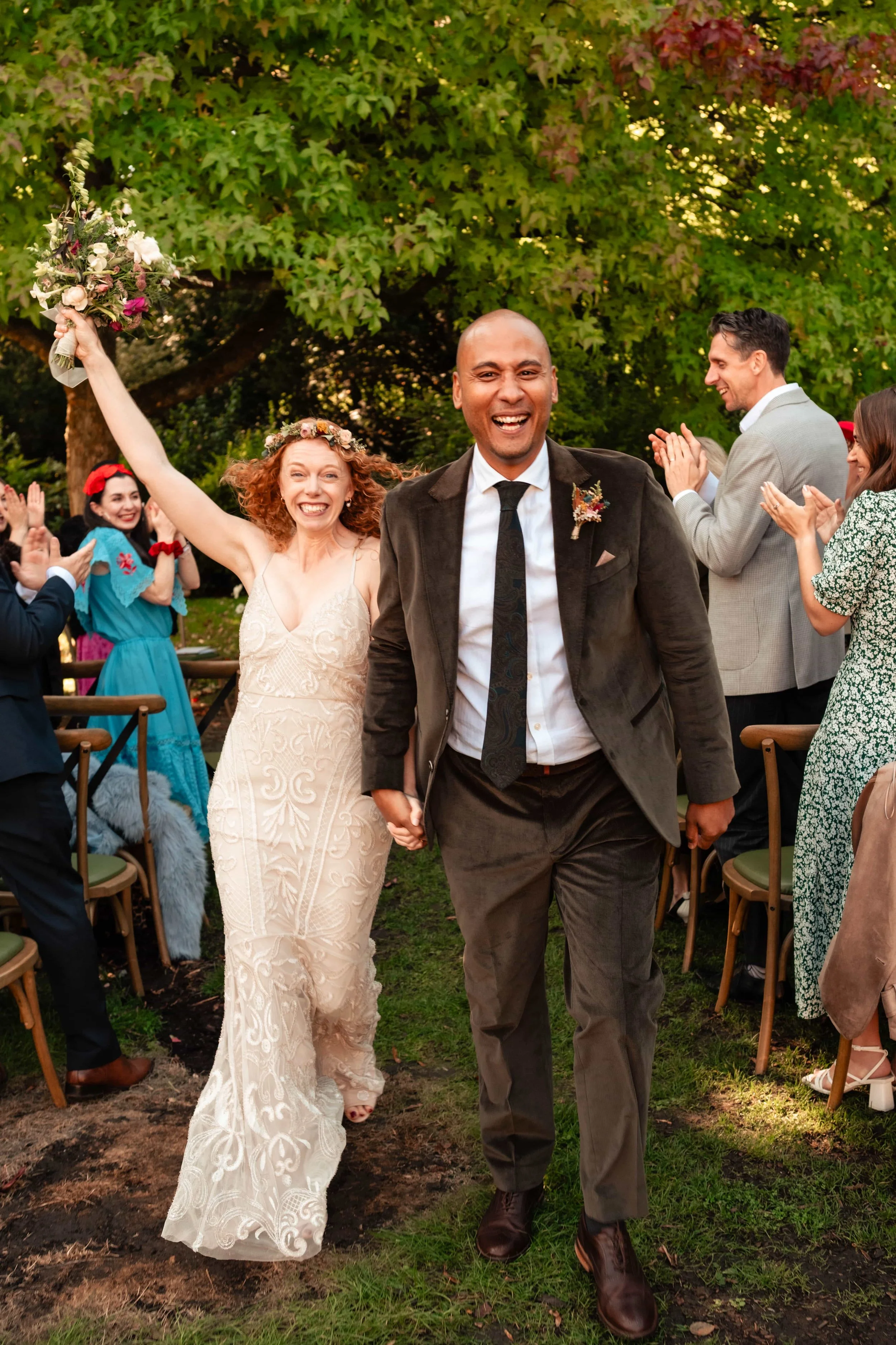 A newly married couple walking down the aisle outdoors, smiling and holding hands, with friends and family celebrating around them. The bride wears a lace wedding dress with a floral headpiece and holds a bouquet, while the groom wears a suit with a 