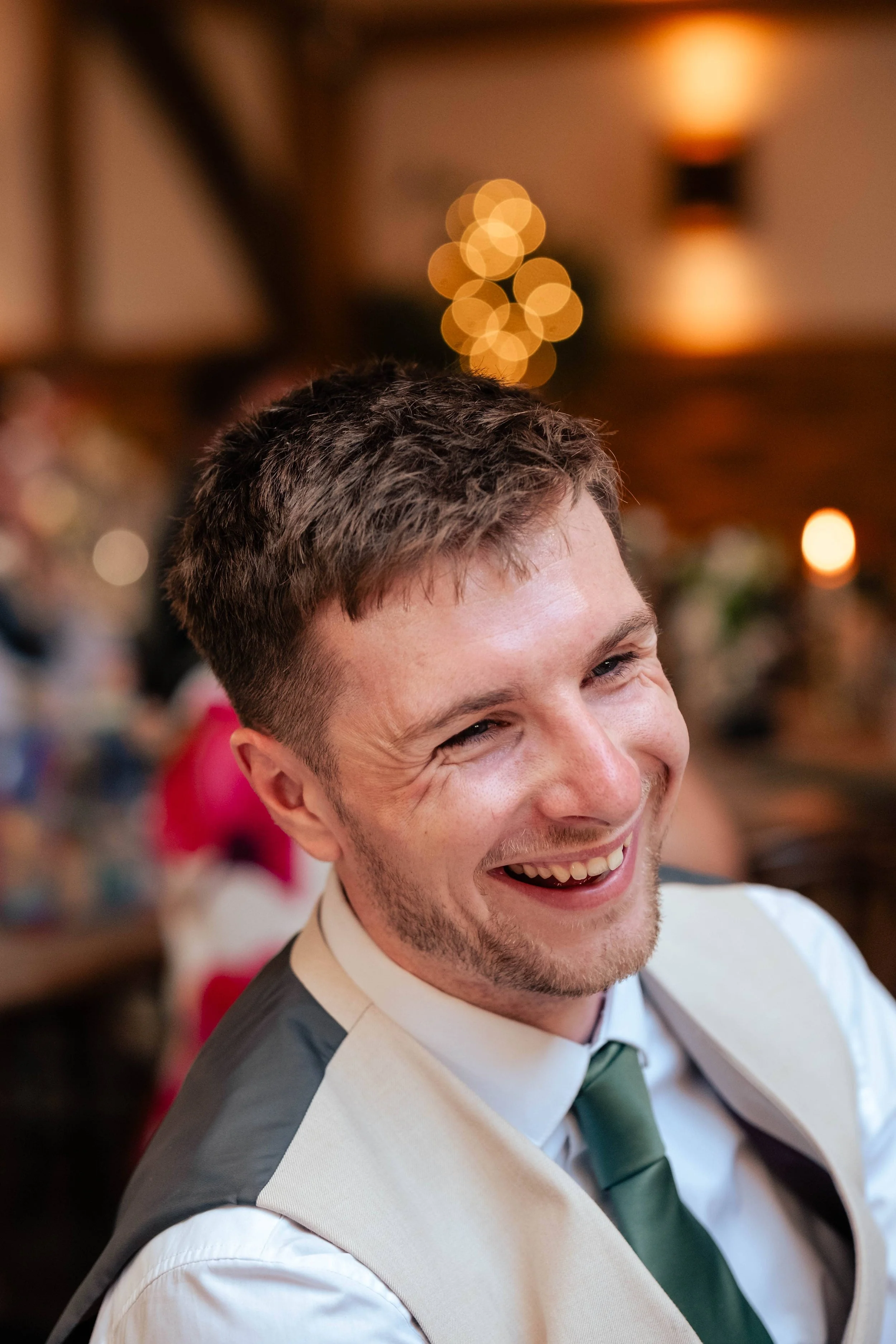A man with short brown hair and beard smiling happily, wearing a white shirt, beige vest, and green tie, in a warmly lit indoor setting with blurred festive or decorative lights in the background.