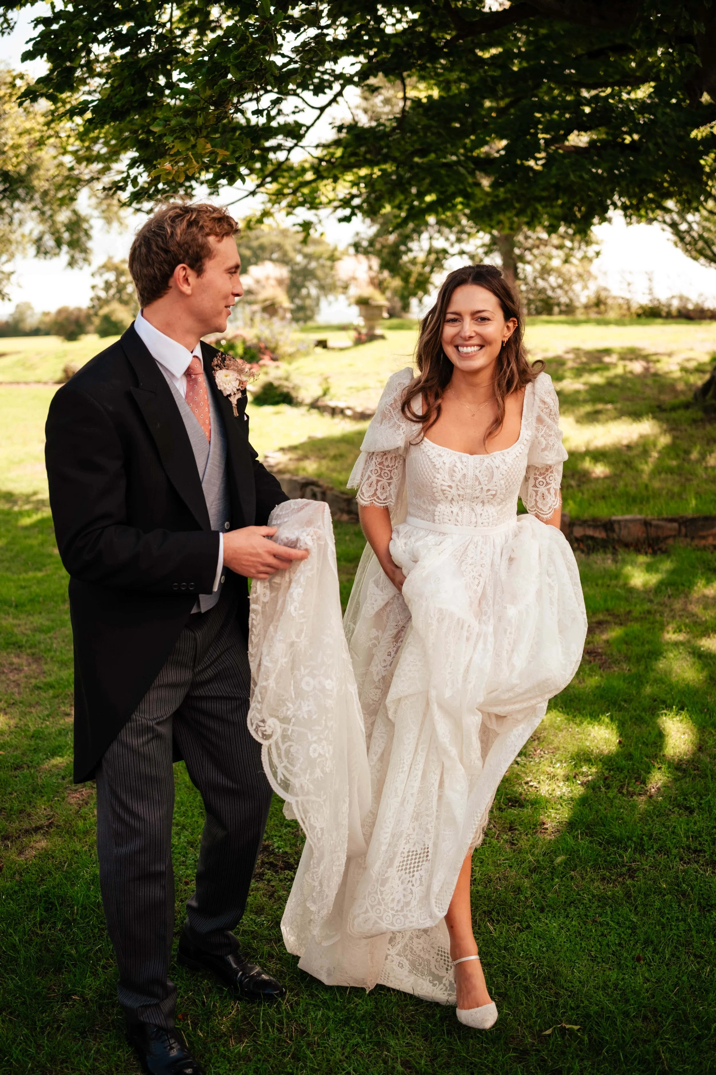 A smiling woman in a white wedding dress holding her dress up while a man in a black suit and striped pants looks at her, standing under a large tree in a grassy outdoor setting.