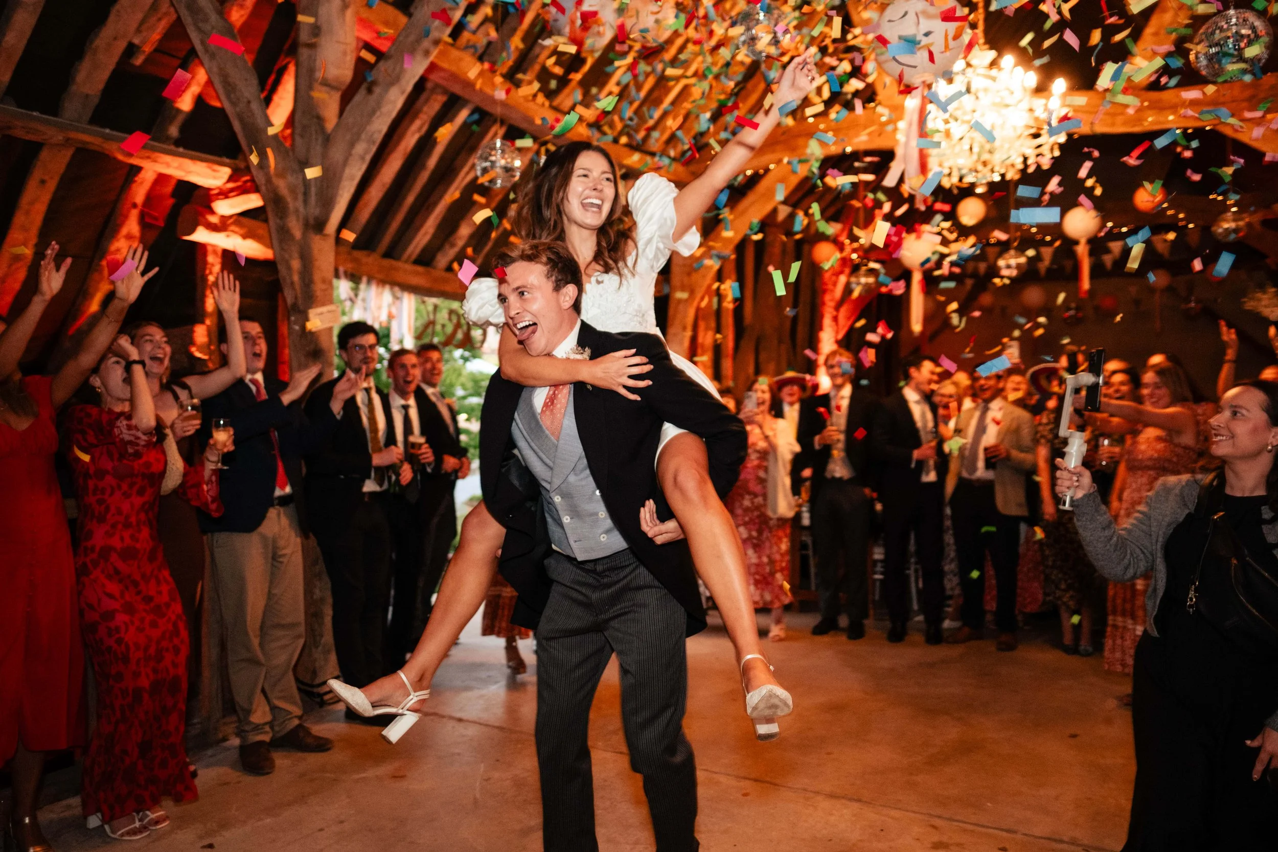 A bride and groom celebrating at their wedding reception with guests cheering and colorful confetti falling, inside a rustic wooden venue.