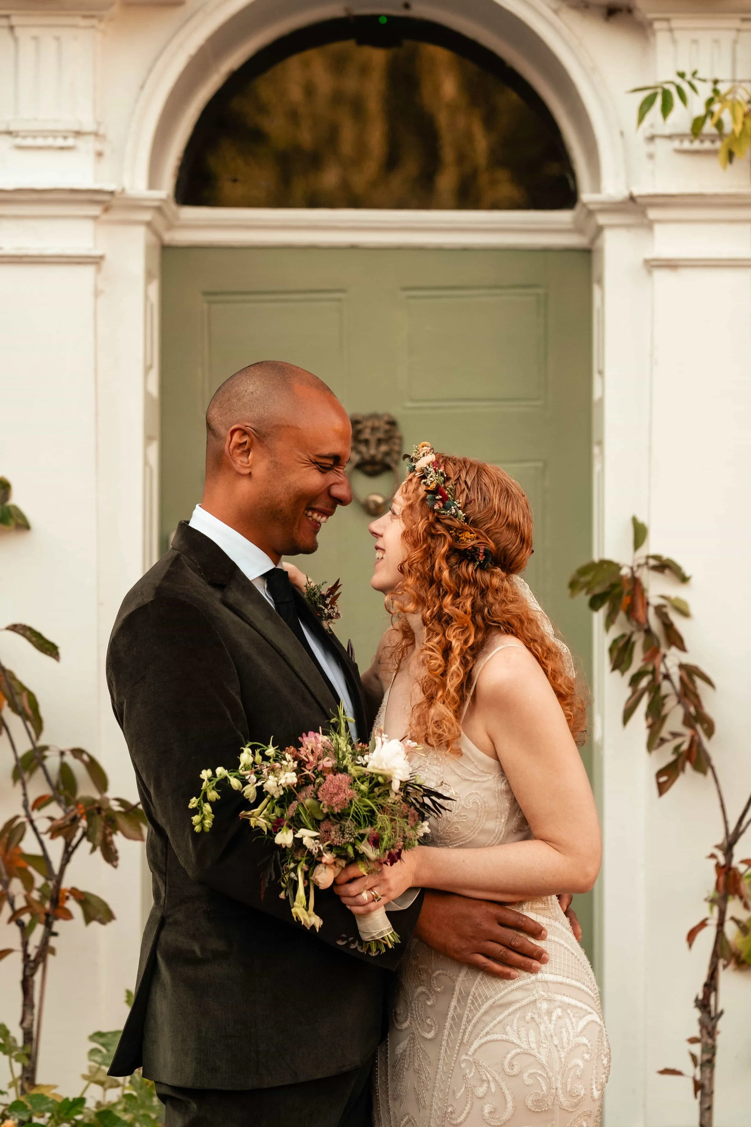 A joyful couple smiling at each other during their wedding, standing in front of a green door with white framing, surrounded by autumnal plants.