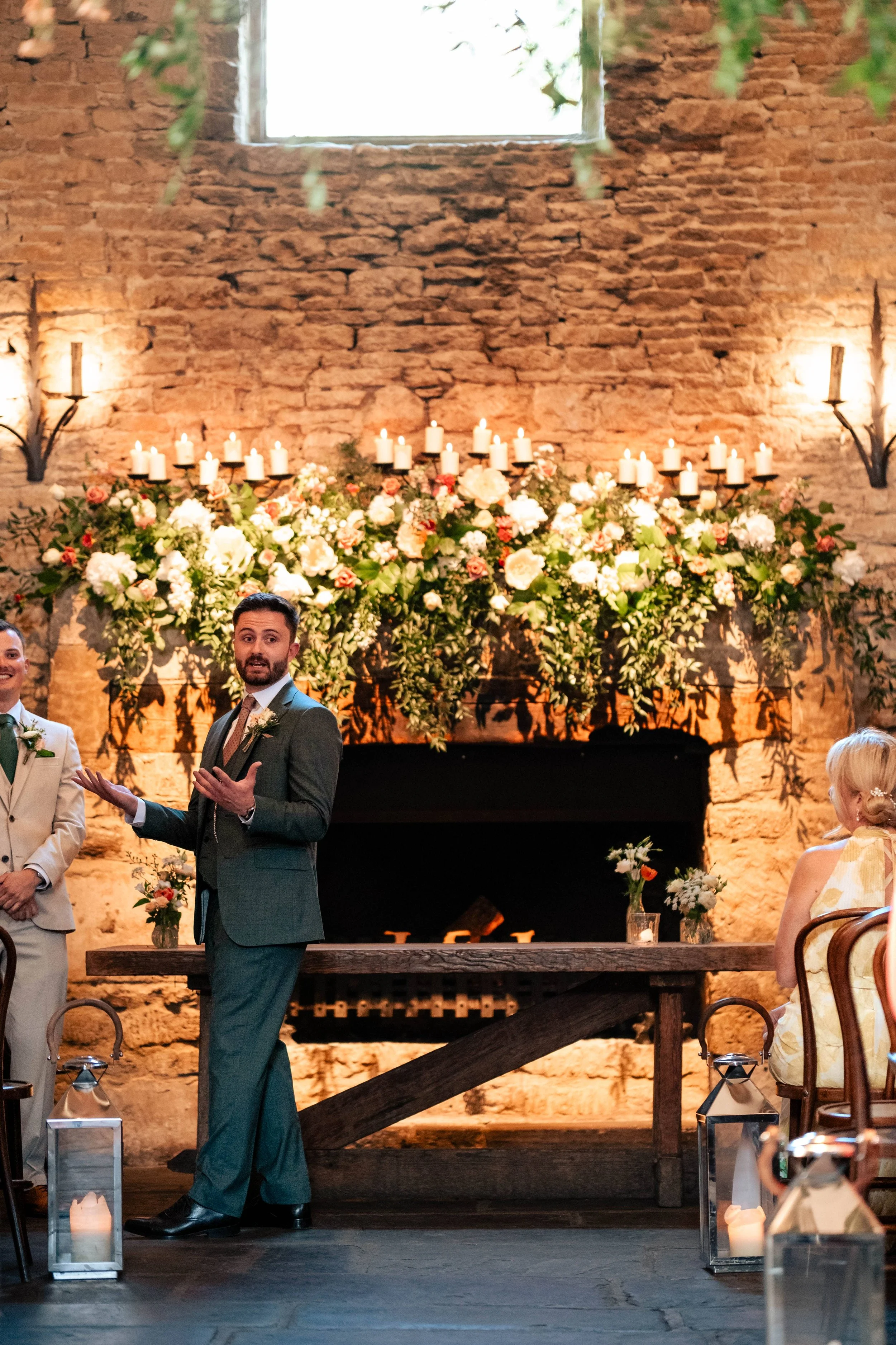 A man giving a speech at a wedding ceremony in front of a decorated brick fireplace with candles and flowers, surrounded by seated guests.