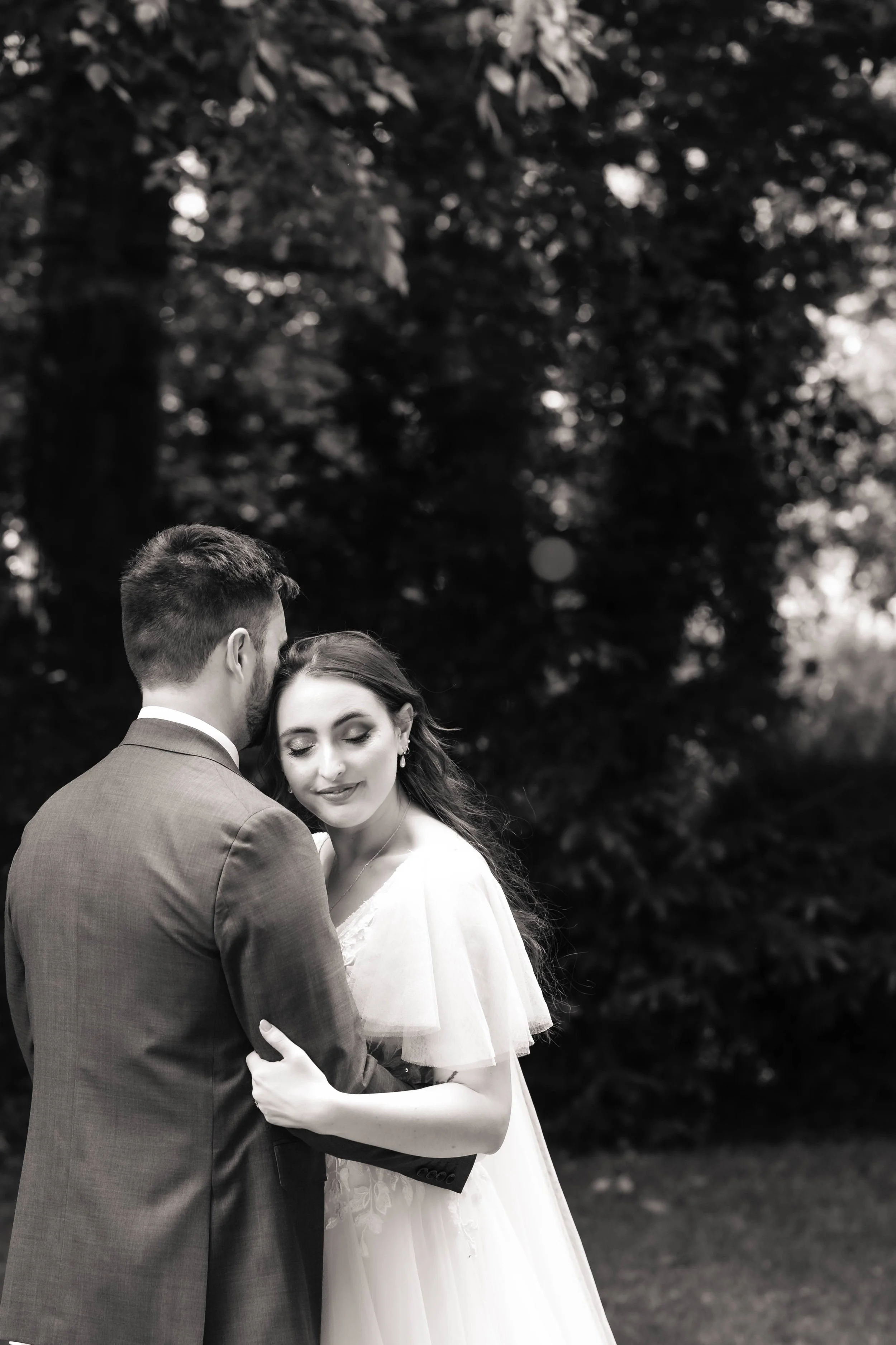 Black and white photo of a bride and groom holding each other outdoors, with trees in the background. The bride has closed eyes and a gentle smile, and the groom's face is turned away from the camera.