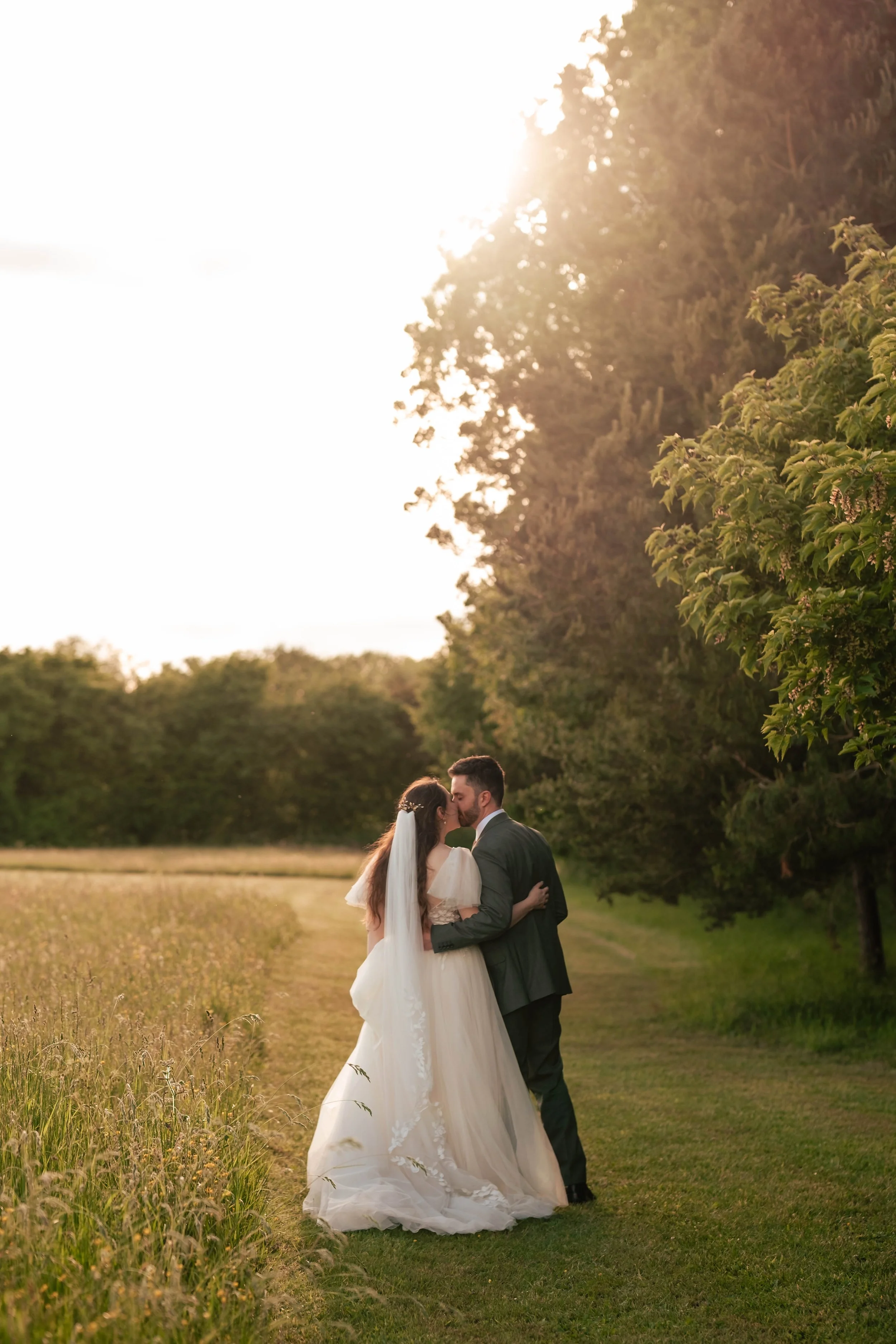 A bride and groom in wedding attire standing and kissing in a grassy field at sunset, surrounded by trees.