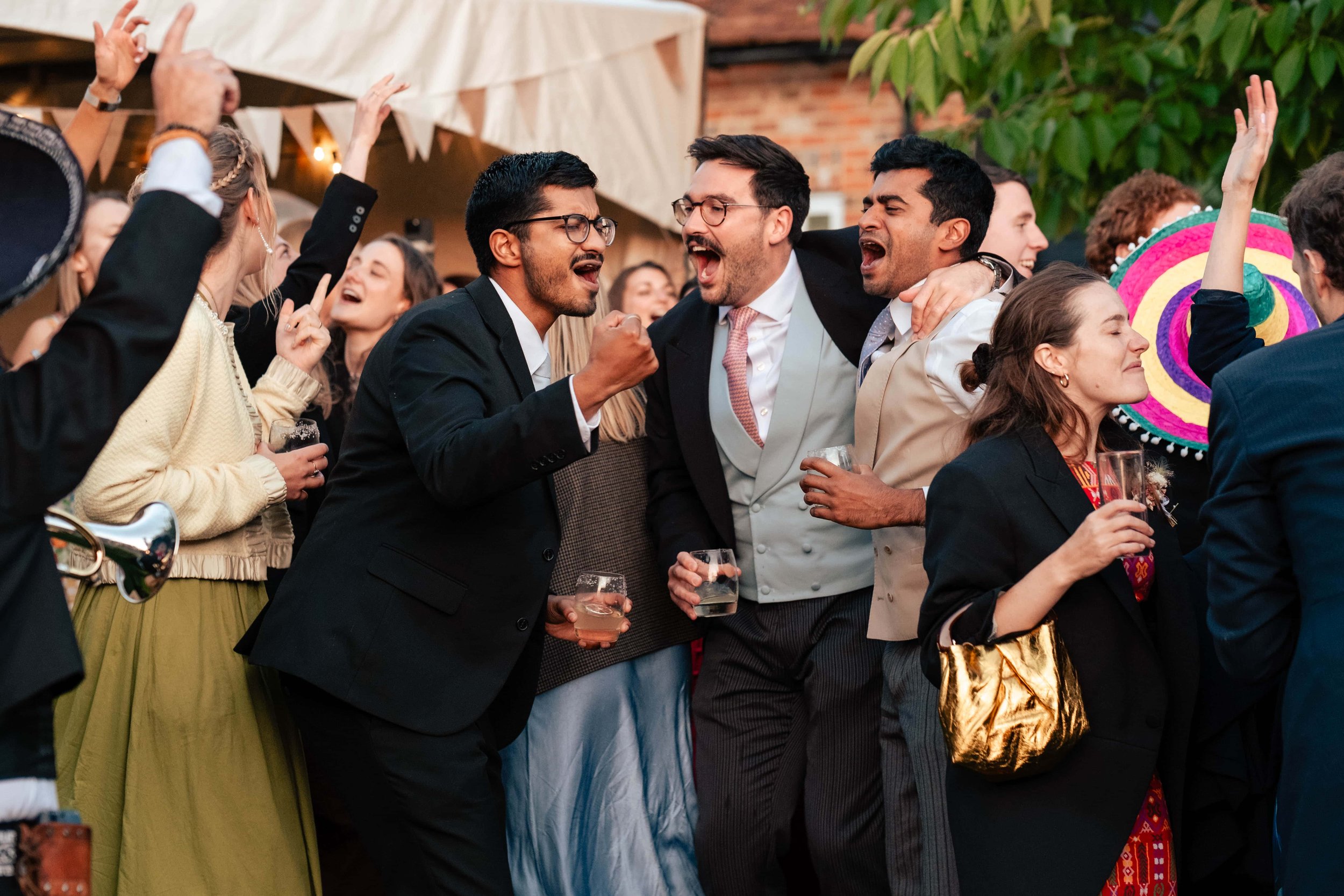 Group of people at a celebration, dressed in formal and festive attire, dancing and having fun outdoors. Some are holding drinks, and a colorful sombrero is visible in the background.