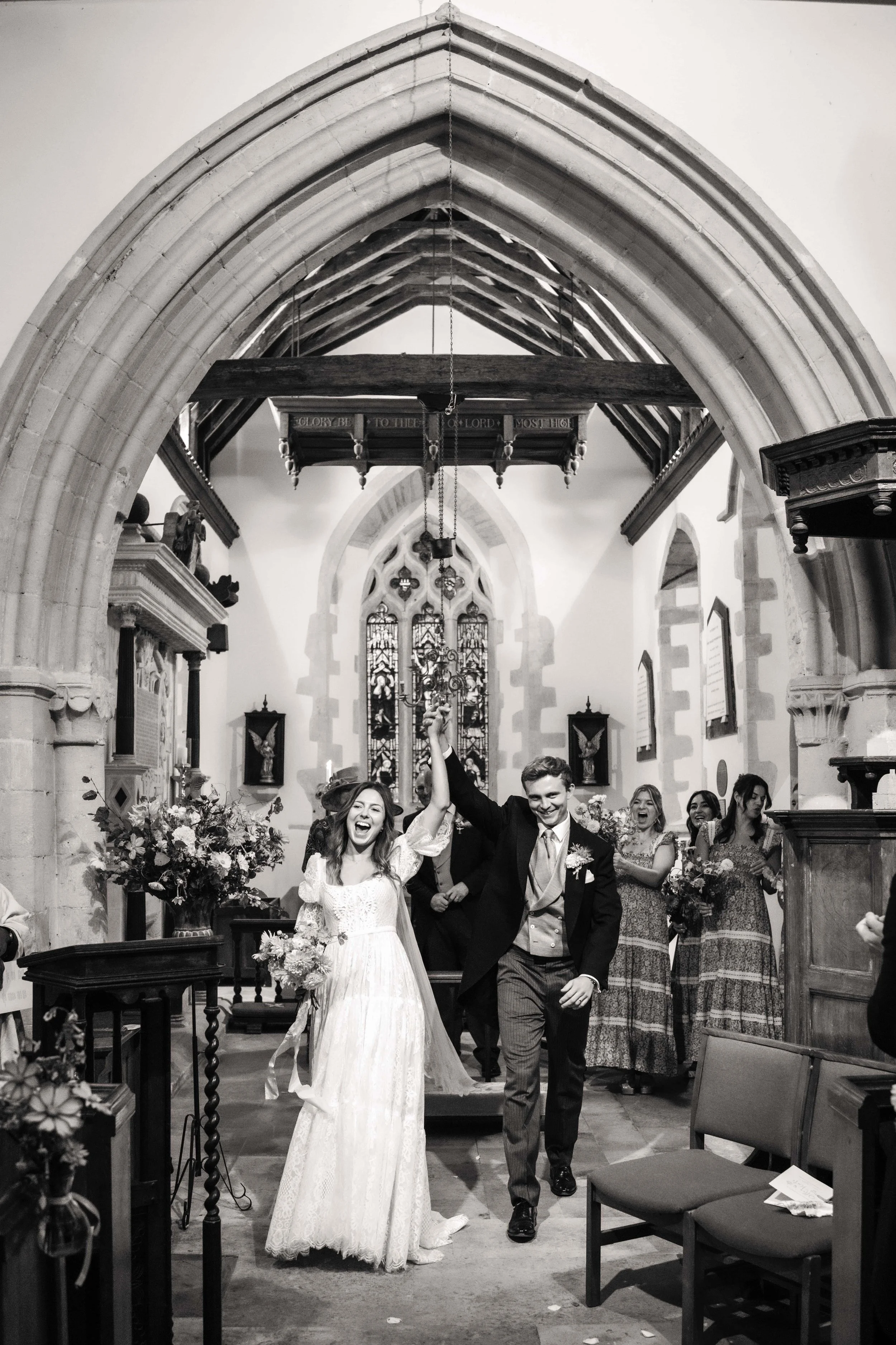 A newly married couple walking down the aisle of a church, holding hands high. The bride is in a white dress, and the groom is in a suit. They are smiling and celebrating as friends and family look on, in a church with stained glass windows and flora