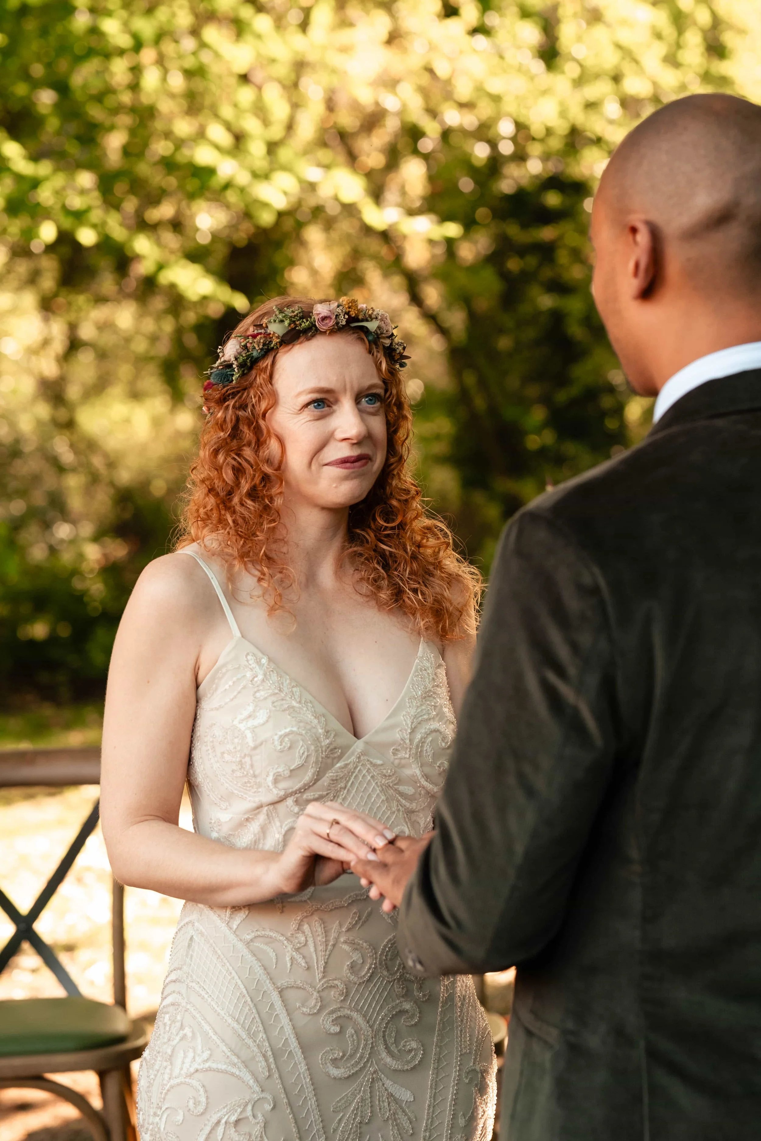 A woman with red curly hair wearing a white dress with intricate embroidery, a floral crown, and holding hands with a man in a dark suit during an outdoor wedding ceremony.
