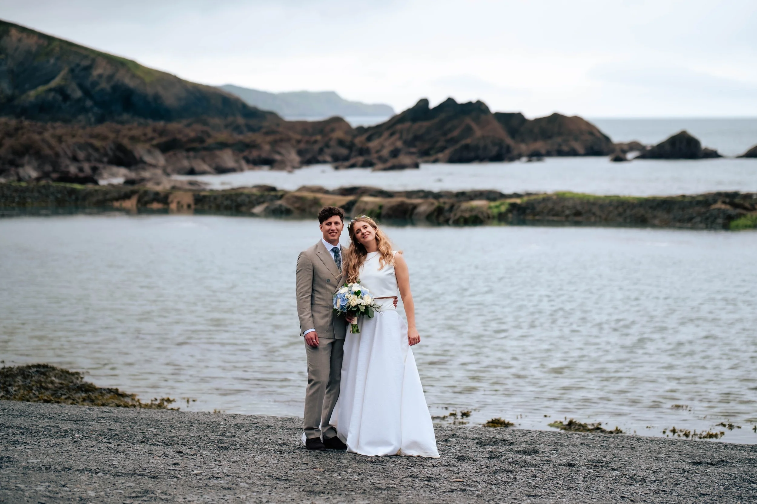 Bride and groom standing together on a beautiful beach, looking towards the camera with the coastline stretching into the distance under an overcast sky.