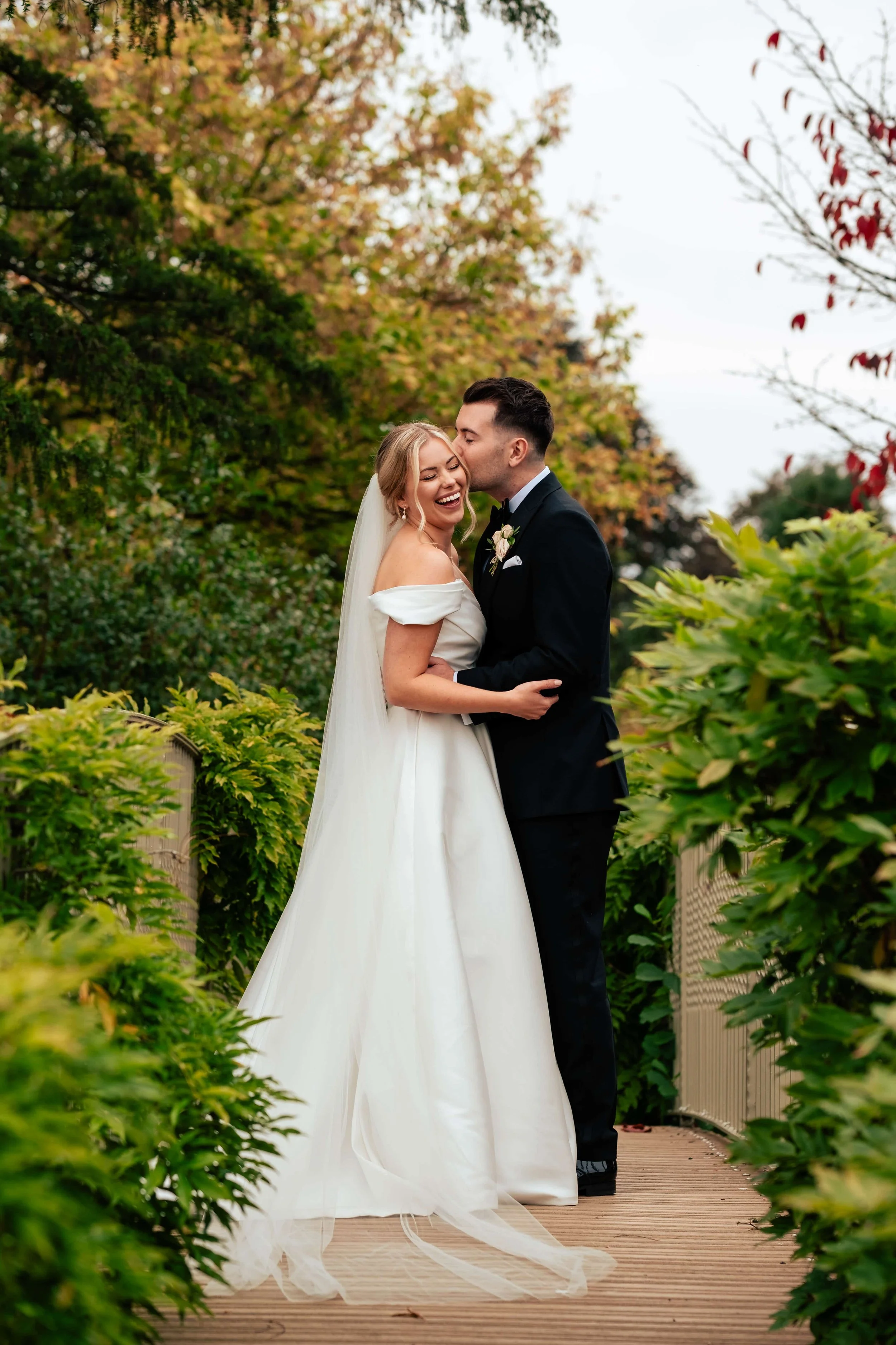 A newlywed couple sharing a joyful moment on a wooden pathway surrounded by lush greenery and colorful trees.