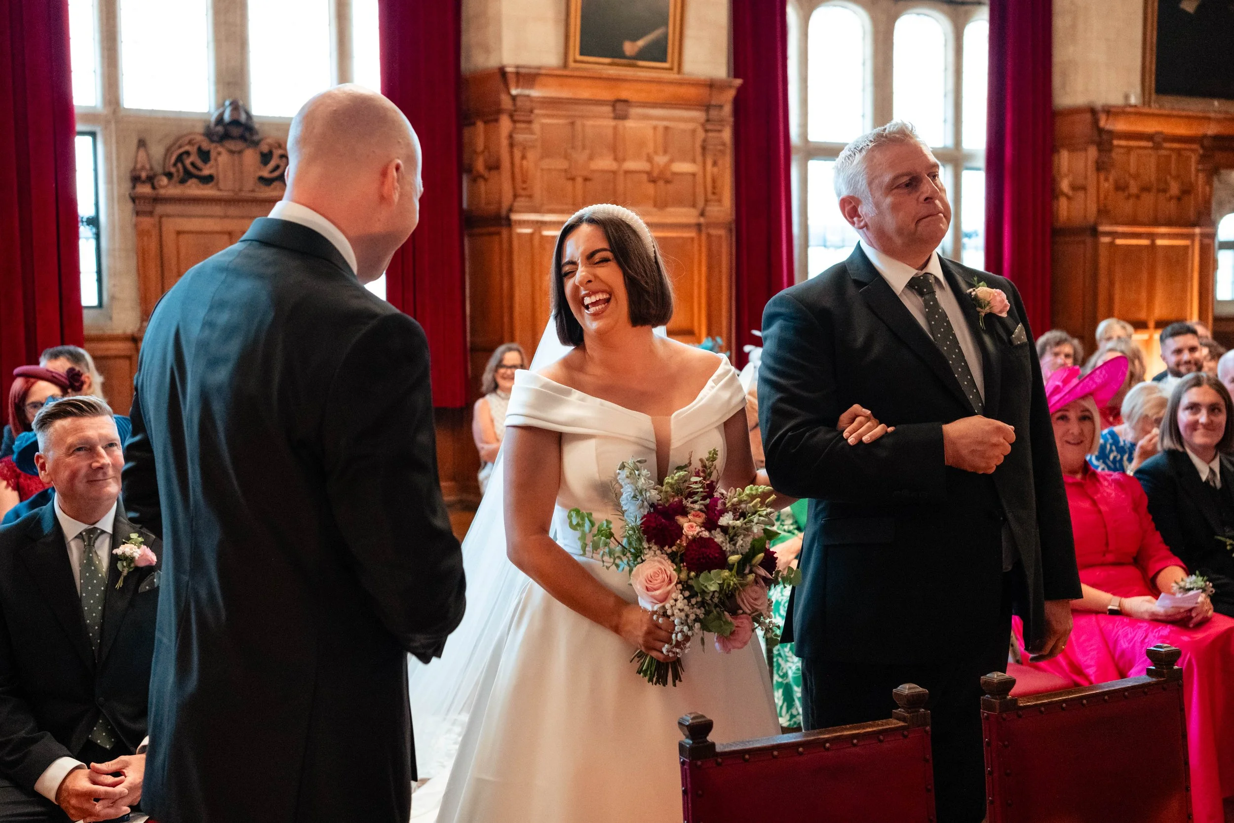 A bride laughing during her wedding ceremony, standing between her groom and an older man, in a wood-paneled room with large windows and red curtains, with seated guests in colorful attire in the background.