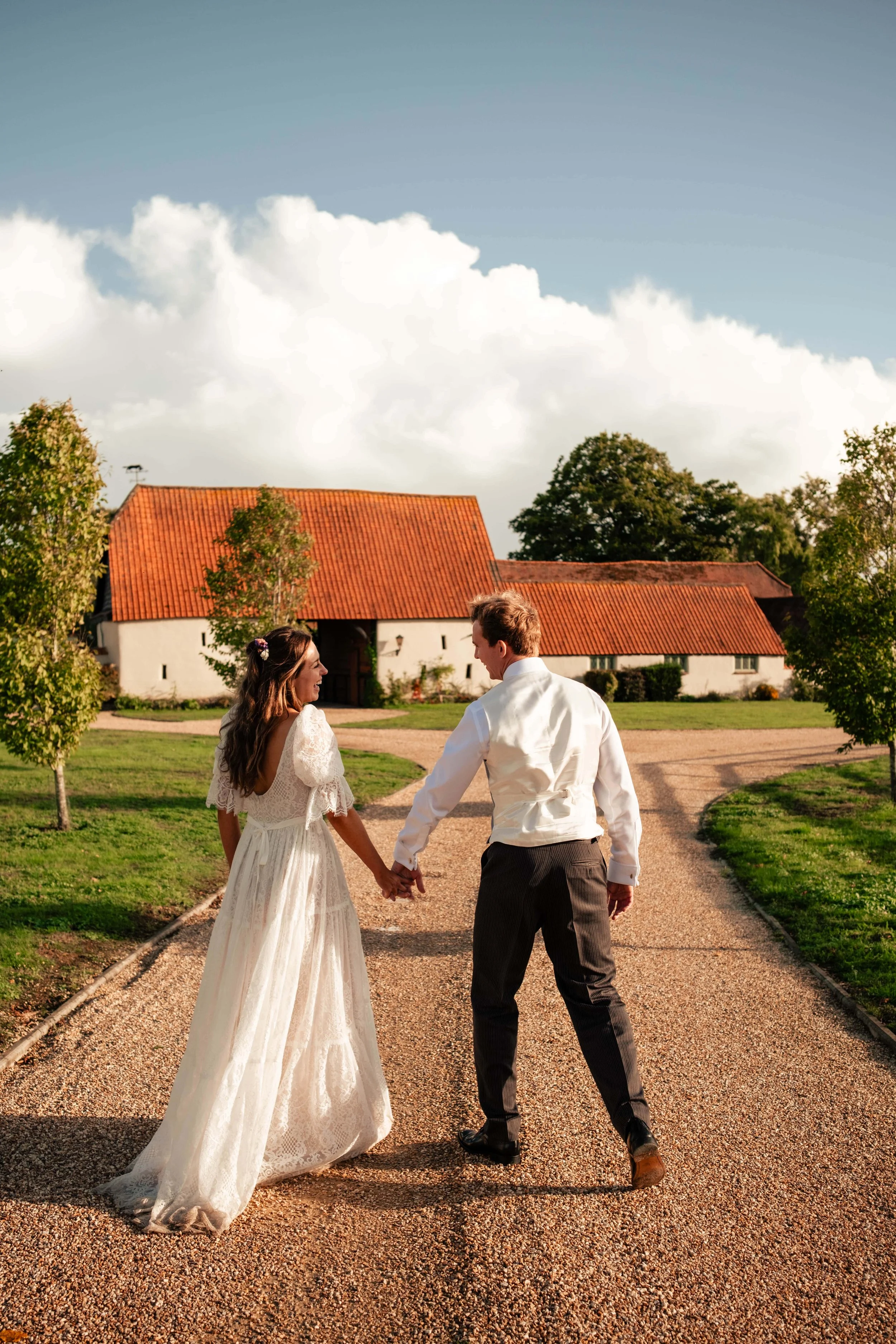 A newlywed couple holding hands and walking towards a rustic barn on a gravel path in a rural setting with green grass, trees, and a partly cloudy sky.
