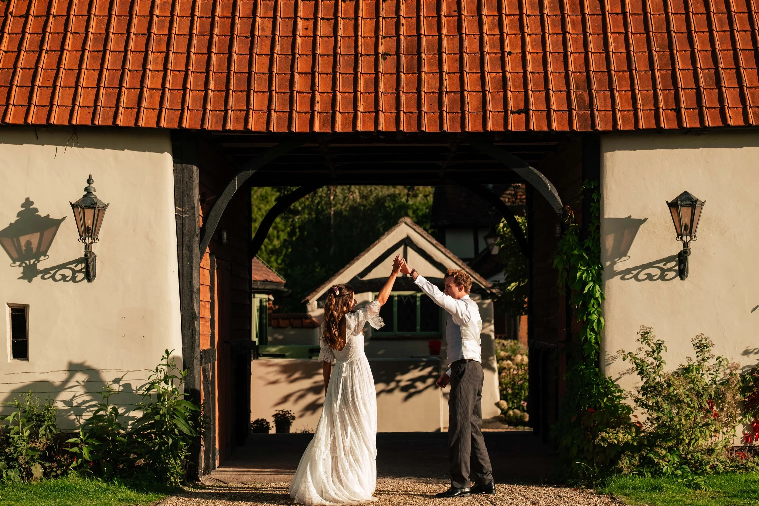 A bride and groom dancing under an archway with a tiled roof, casting shadows on the walls, as sunlight illuminates the scene.