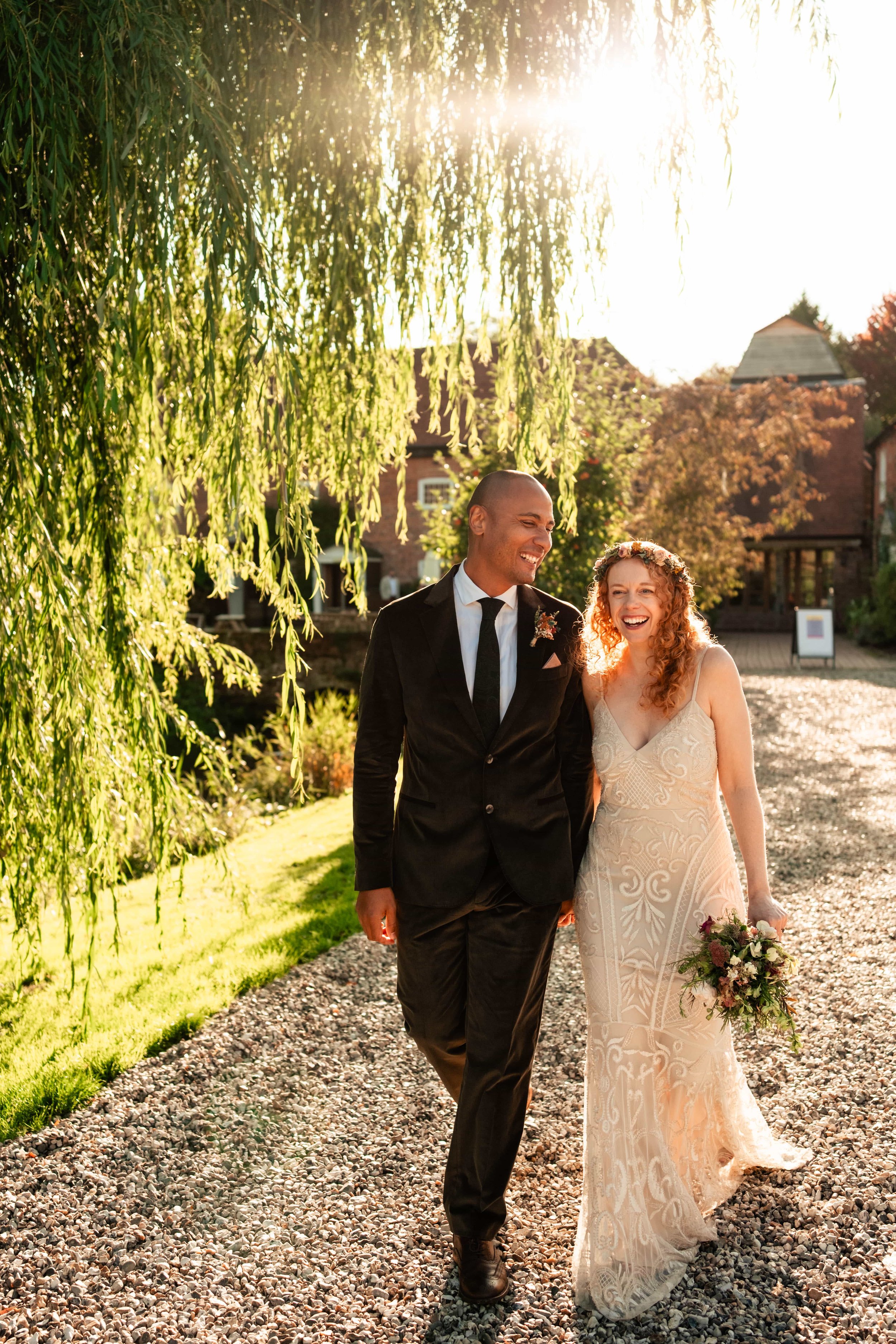 A smiling bride and groom walking outdoors on a gravel path, with the bride holding a bouquet of flowers and wearing a floral crown, as the sun sets behind them and creates a warm glow.