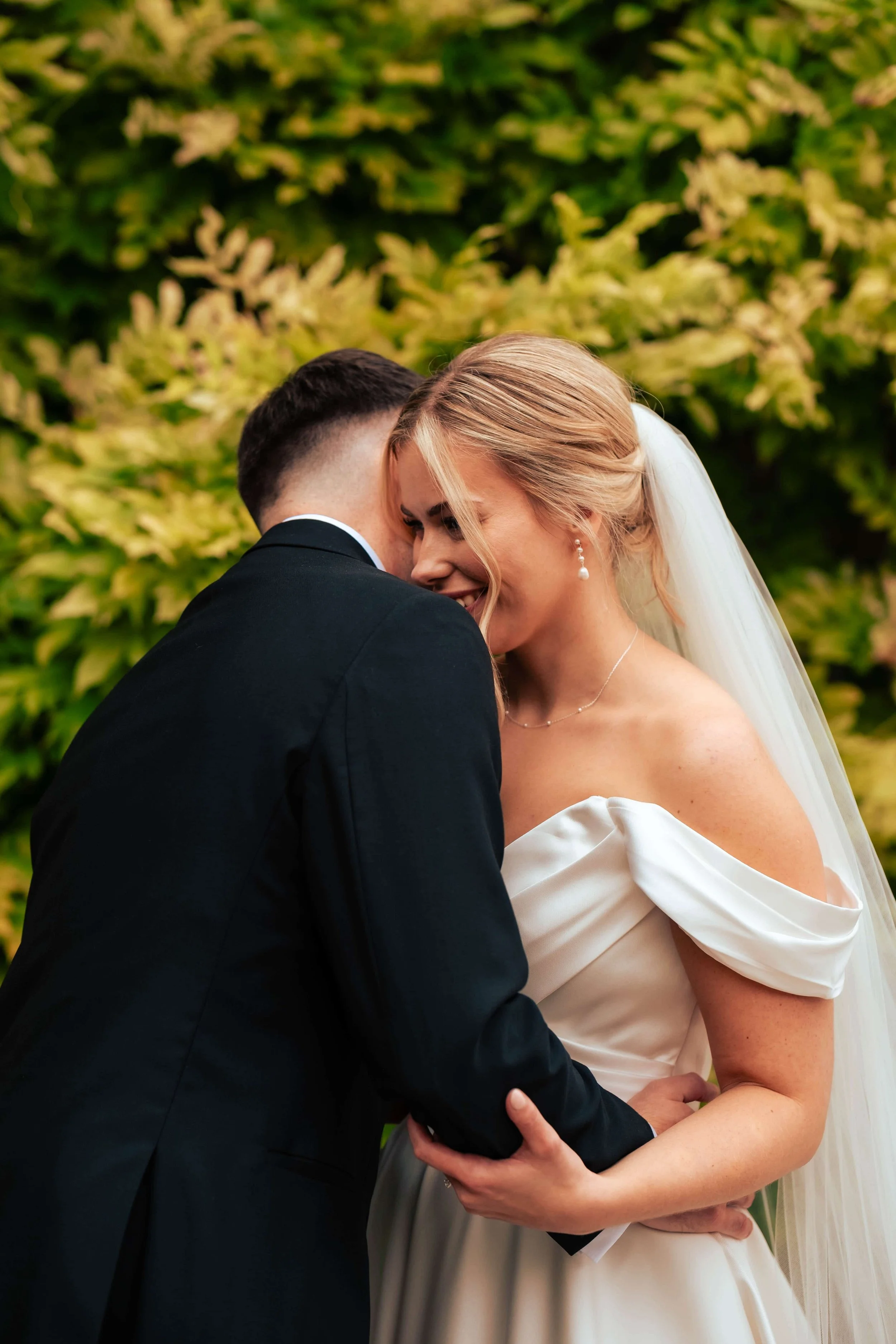 A bride and groom sharing an intimate moment, embracing each other outdoors against a backdrop of green foliage.