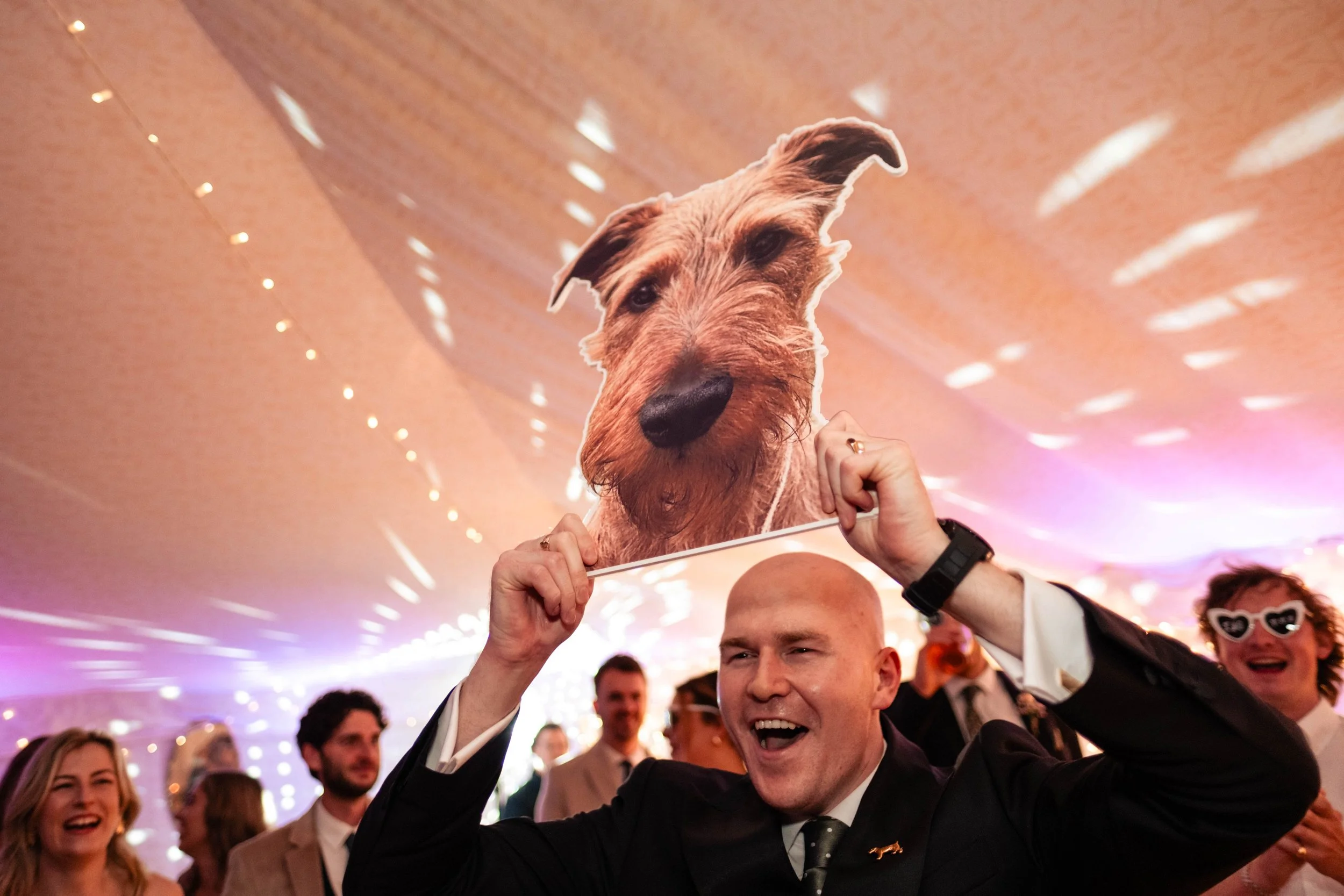Man holding a dog face mask at a party or celebration, with other people in the background.