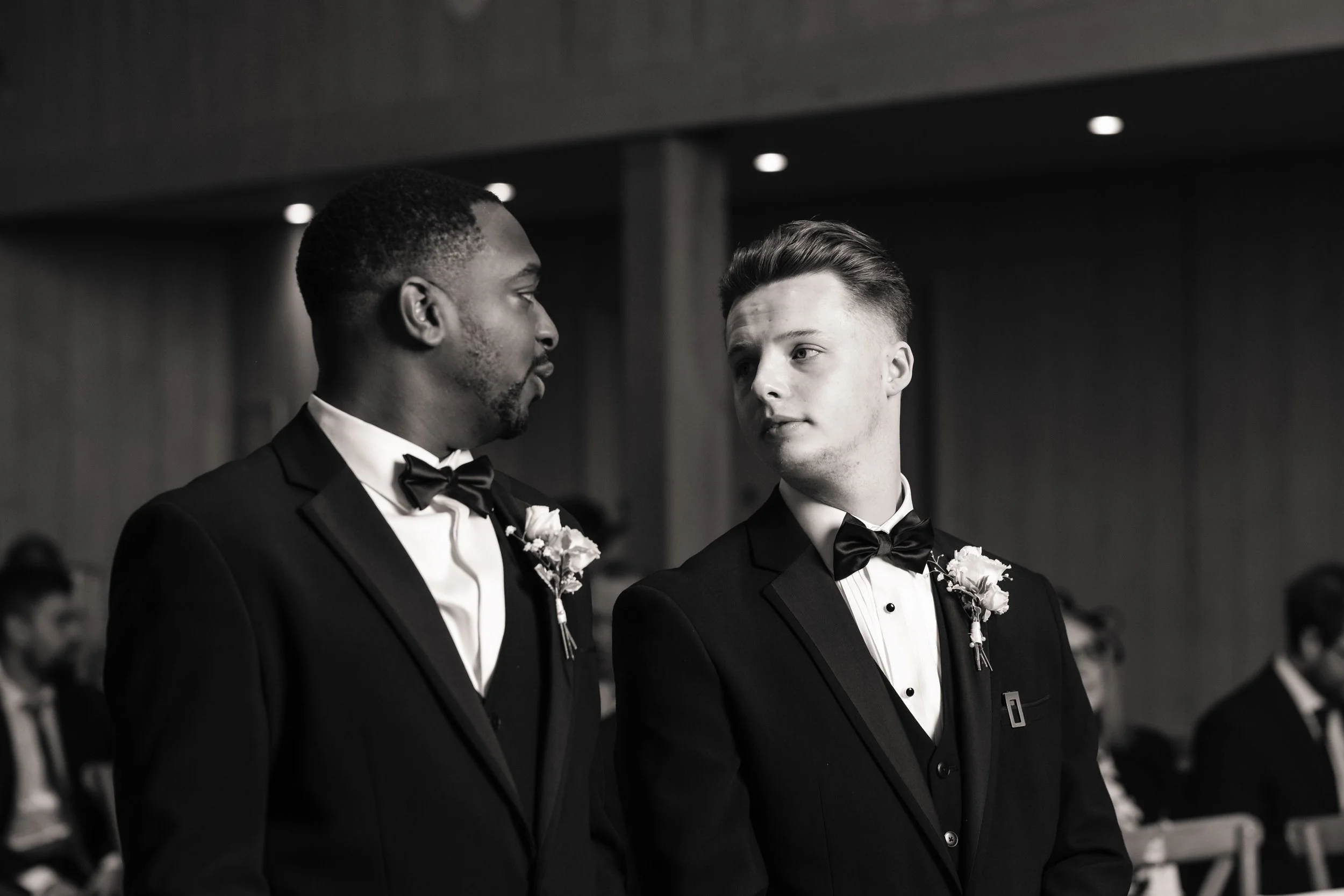 The groom and his best man in tuxedos with bow ties and boutonnières, standing face-to-face waiting for the brides arrival, in a black-and-white photo.