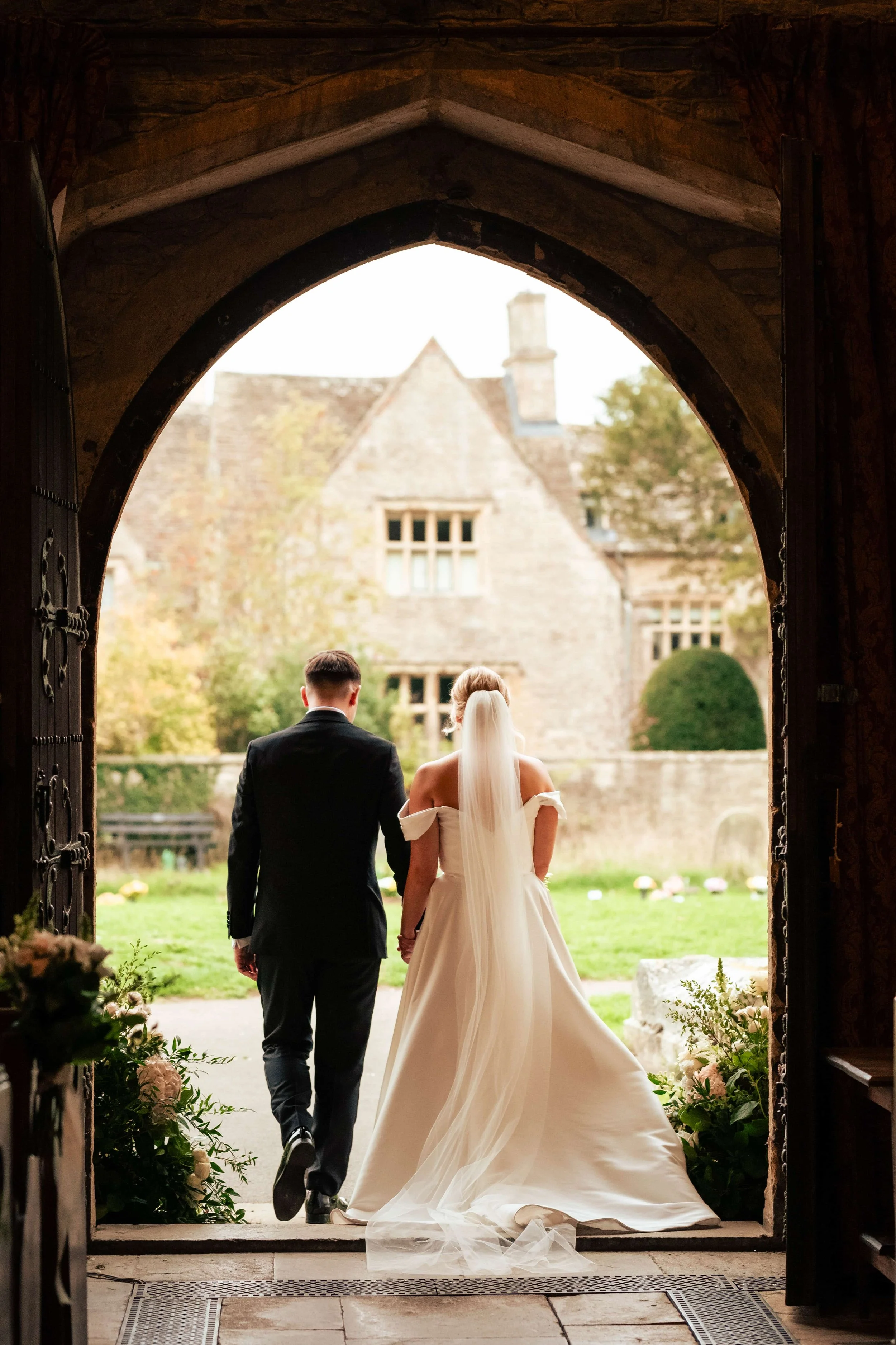 A bride and groom walking out of a stone archway of a church or historic building, with a lush green lawn and historic stone buildings in the background, during a wedding ceremony.