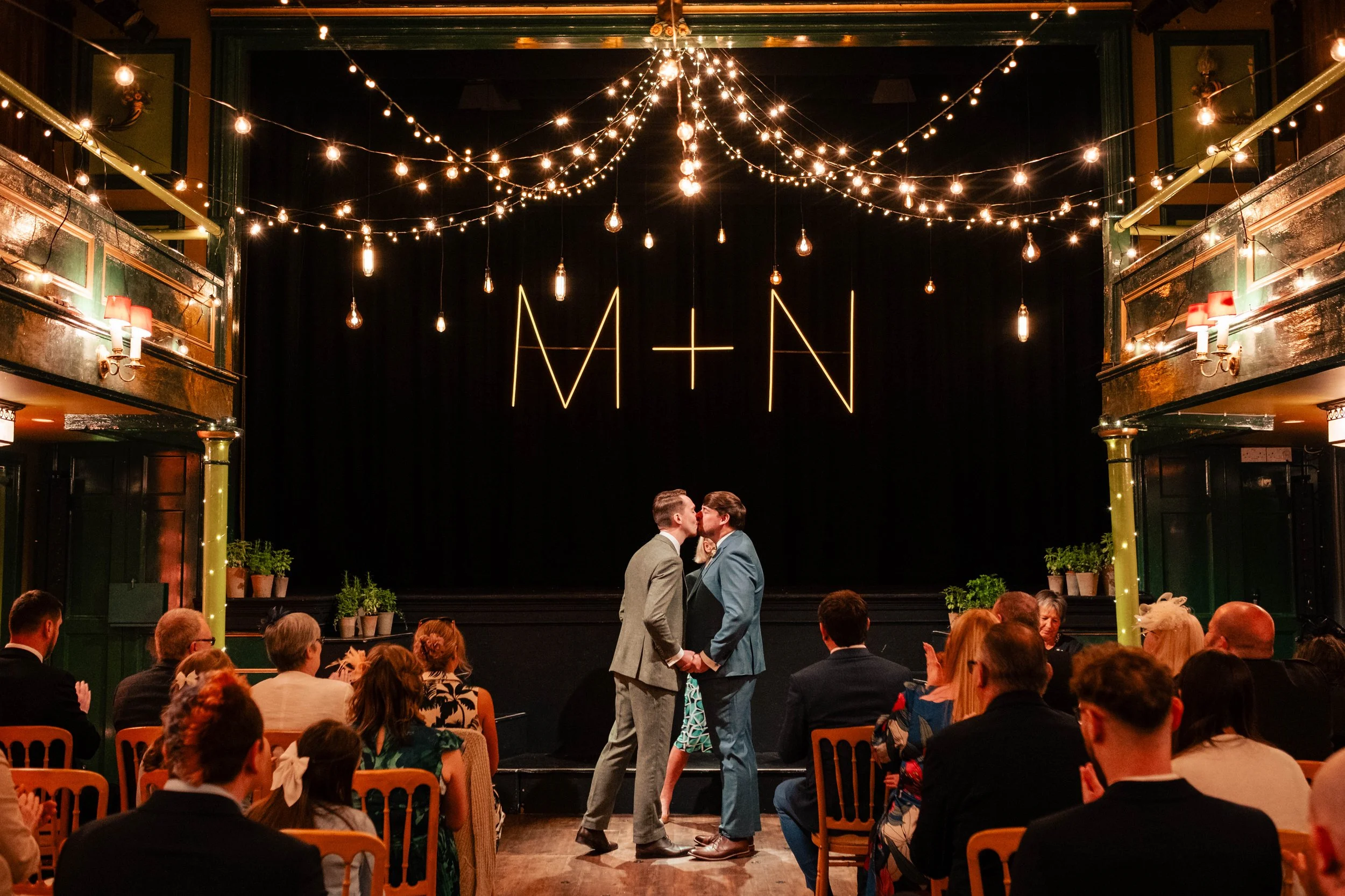A same-sex couple shares a kiss on stage at their wedding ceremony in front of their guests, with decorative string lights and a neon sign displaying 'M + N' in the background.