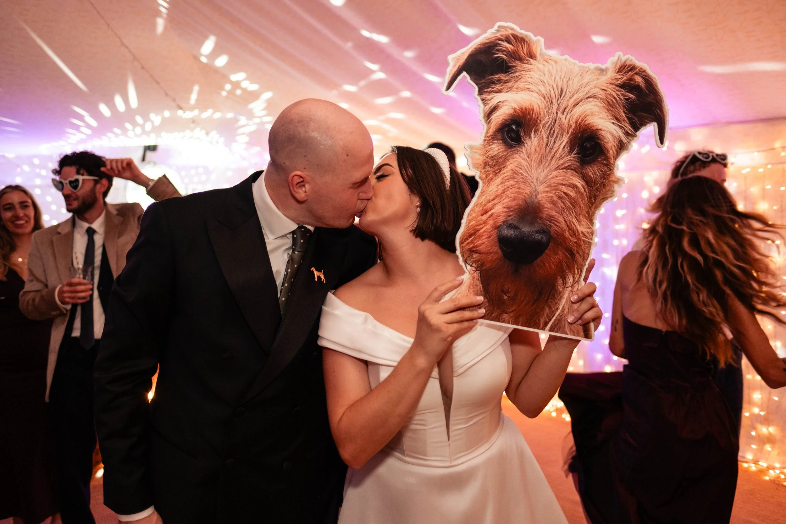 Couple kissing at wedding reception, woman holding a dog face mask, guests dancing in background with colorful lights.