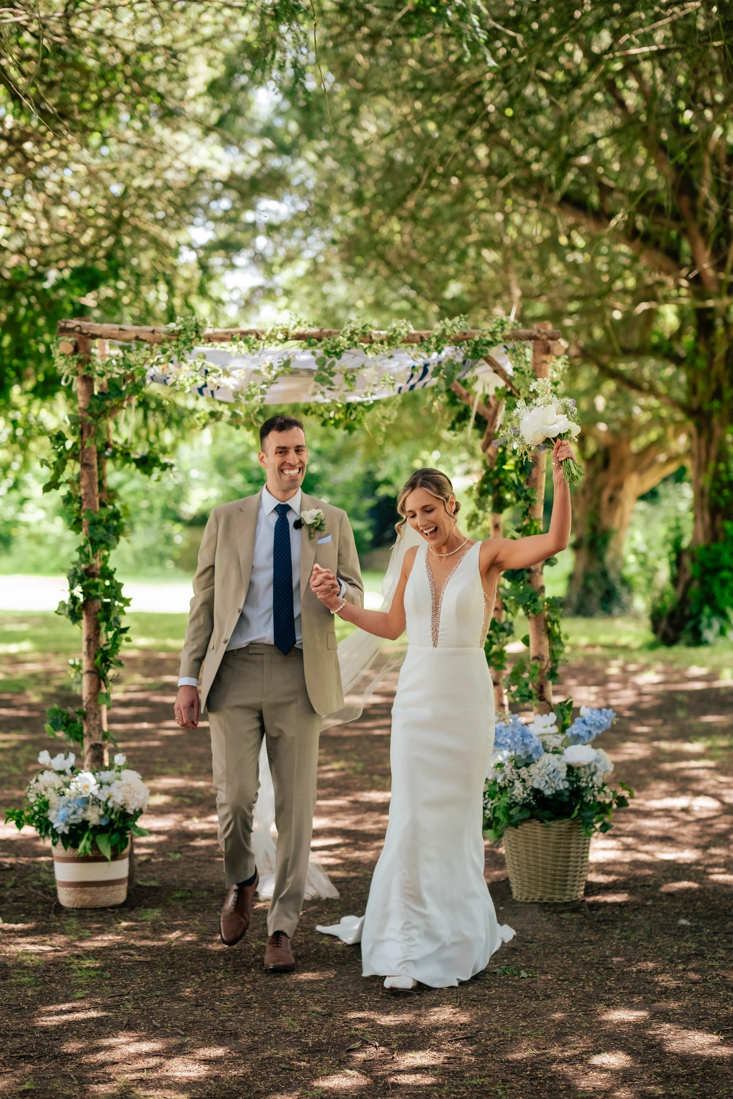 Bride and groom walking down the aisle after their wedding ceremony, with the bride raising her flowers in the air and trees surrounding the outdoor ceremony.