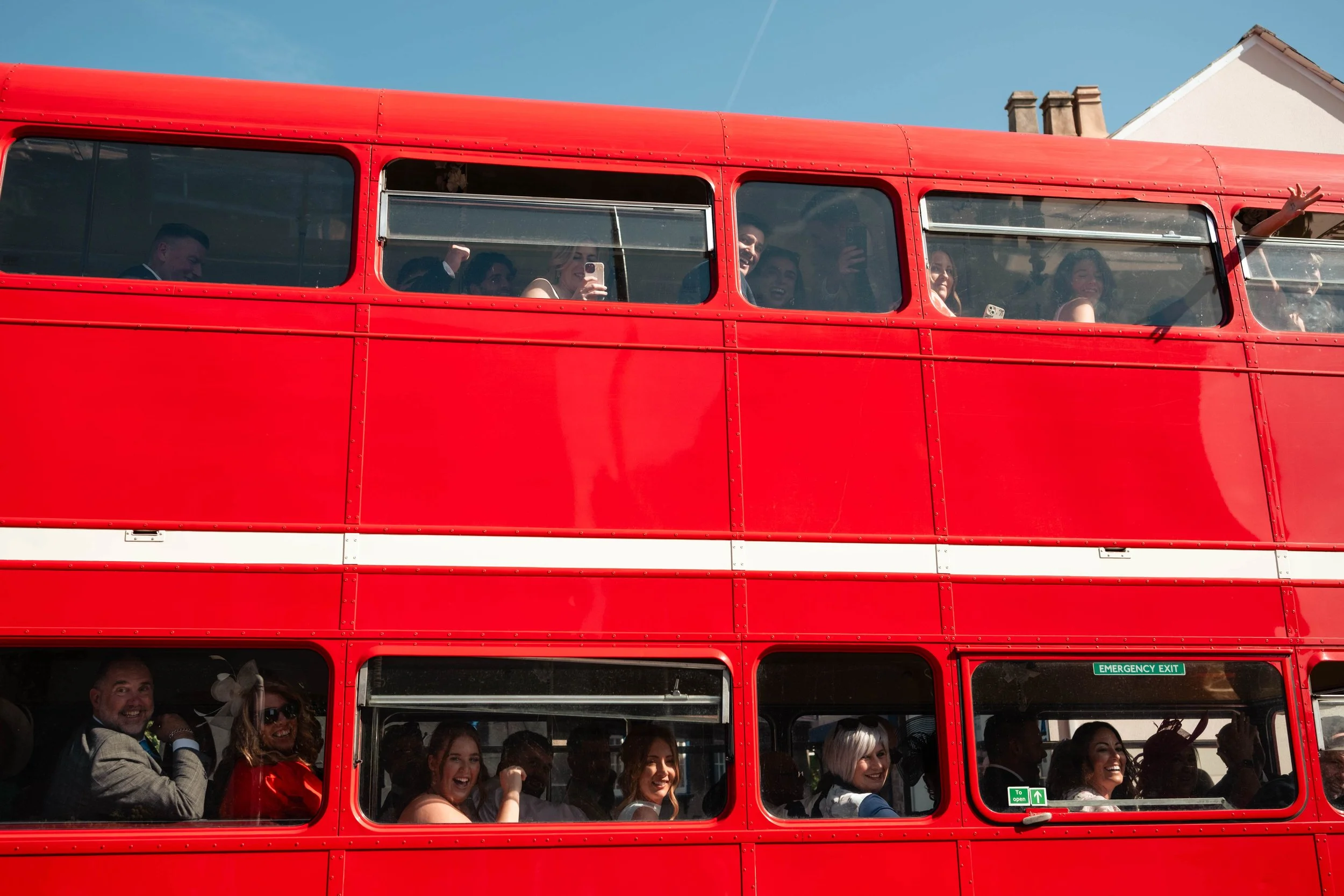 Red double-decker bus filled with happy and smiling people, some taking photos and waving, set against a clear blue sky.