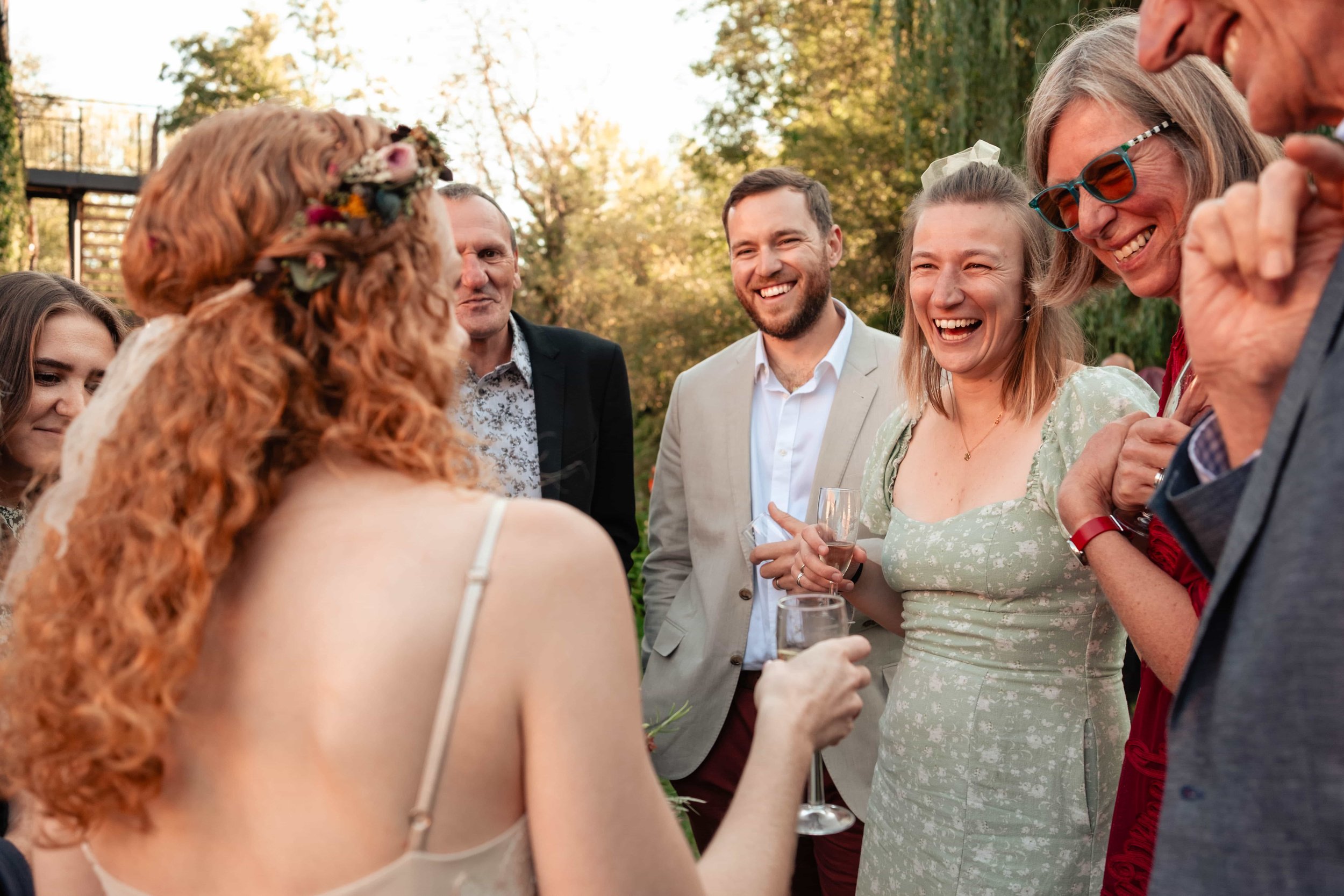 People at an outdoor celebration, with a woman with red curly hair and a floral headband in focus, holding a glass of wine and smiling, surrounded by friends in formal attire
