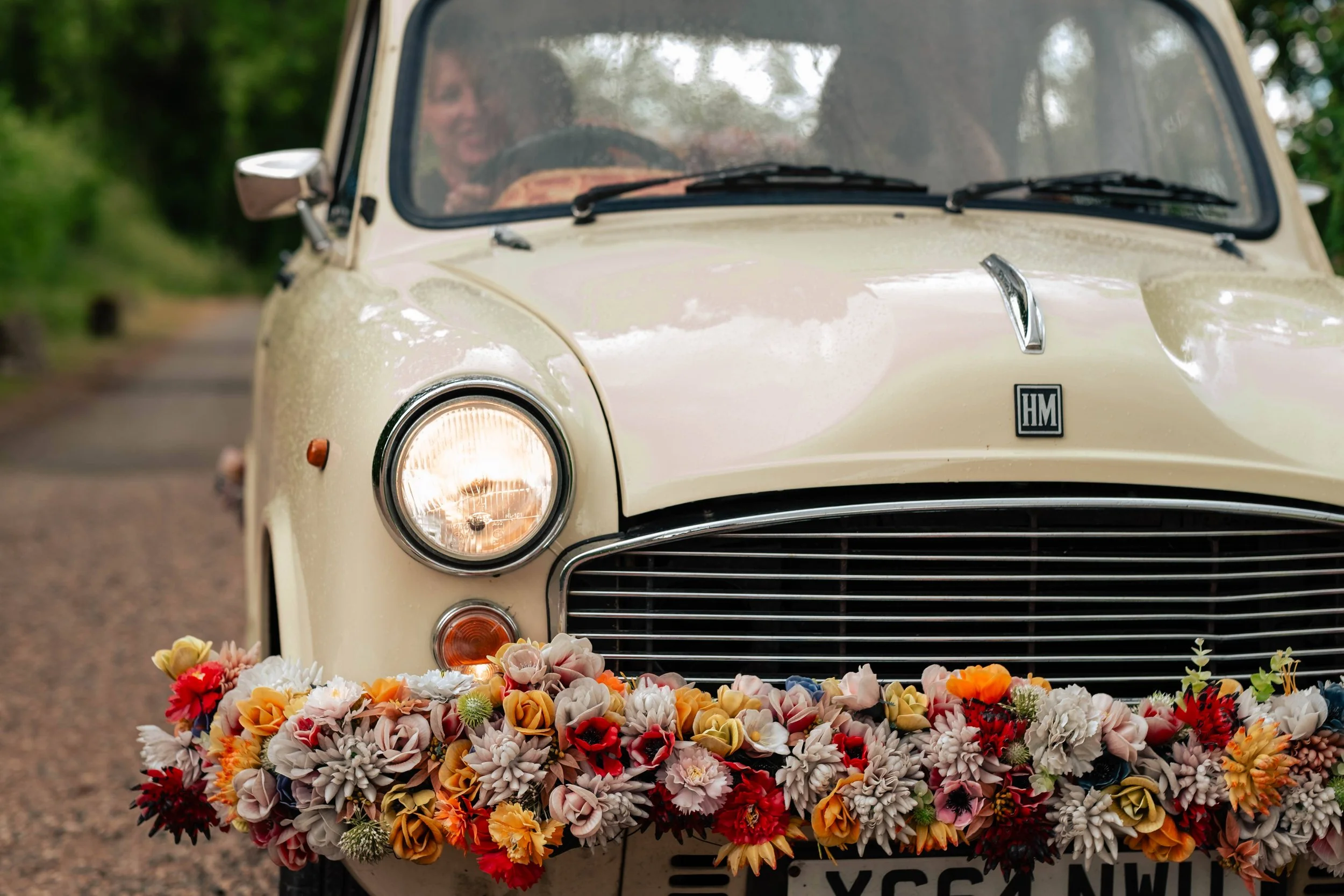 A vintage cream-colored car decorated with a colorful flower arrangement on the front bumper, parked on a rural dirt road with trees in the background.