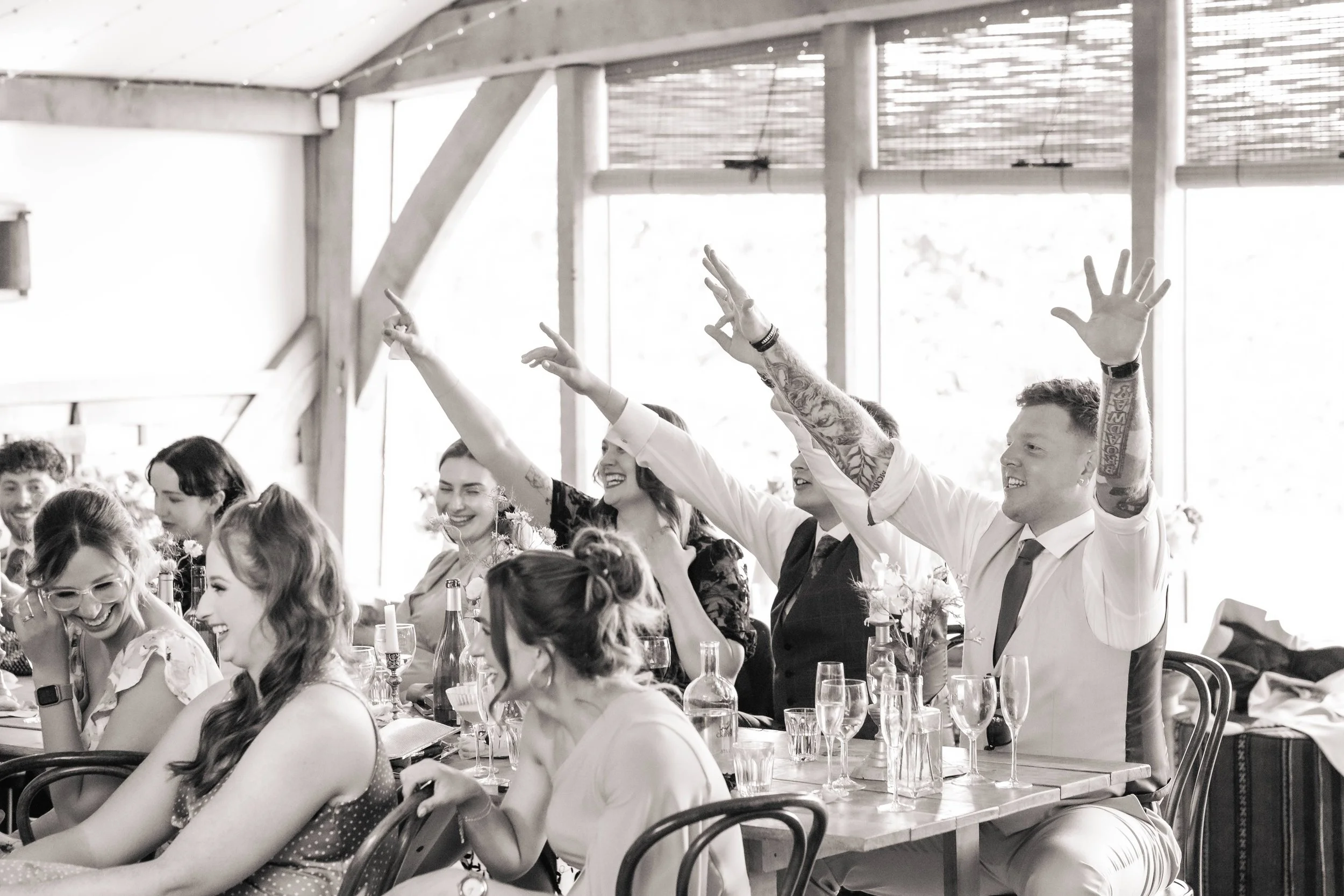 People at a wedding reception seated at long tables, smiling and raising their hands in celebration.
