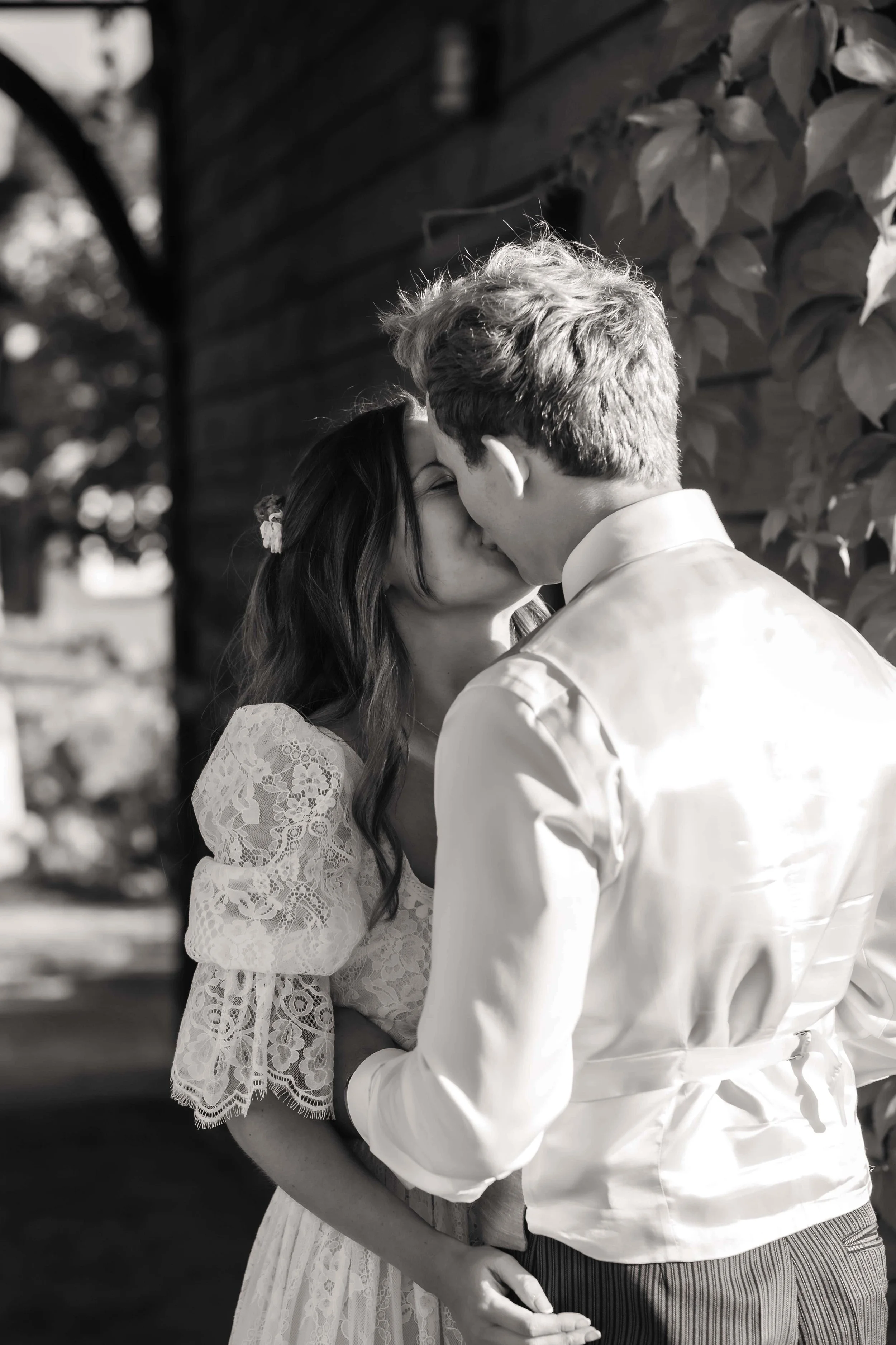 Black and white photo of a couple kissing outdoors, with the woman wearing a lace dress and the man in a light-colored jacket.