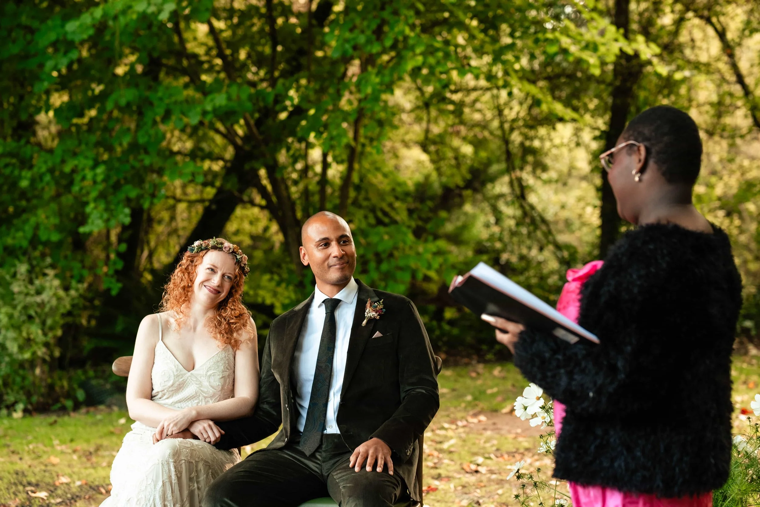 A wedding ceremony outdoors in a wooded area, featuring a woman in a white dress with red curly hair and a floral headband, a man in a dark suit, and an officiant woman in black with pink accents holding a book.