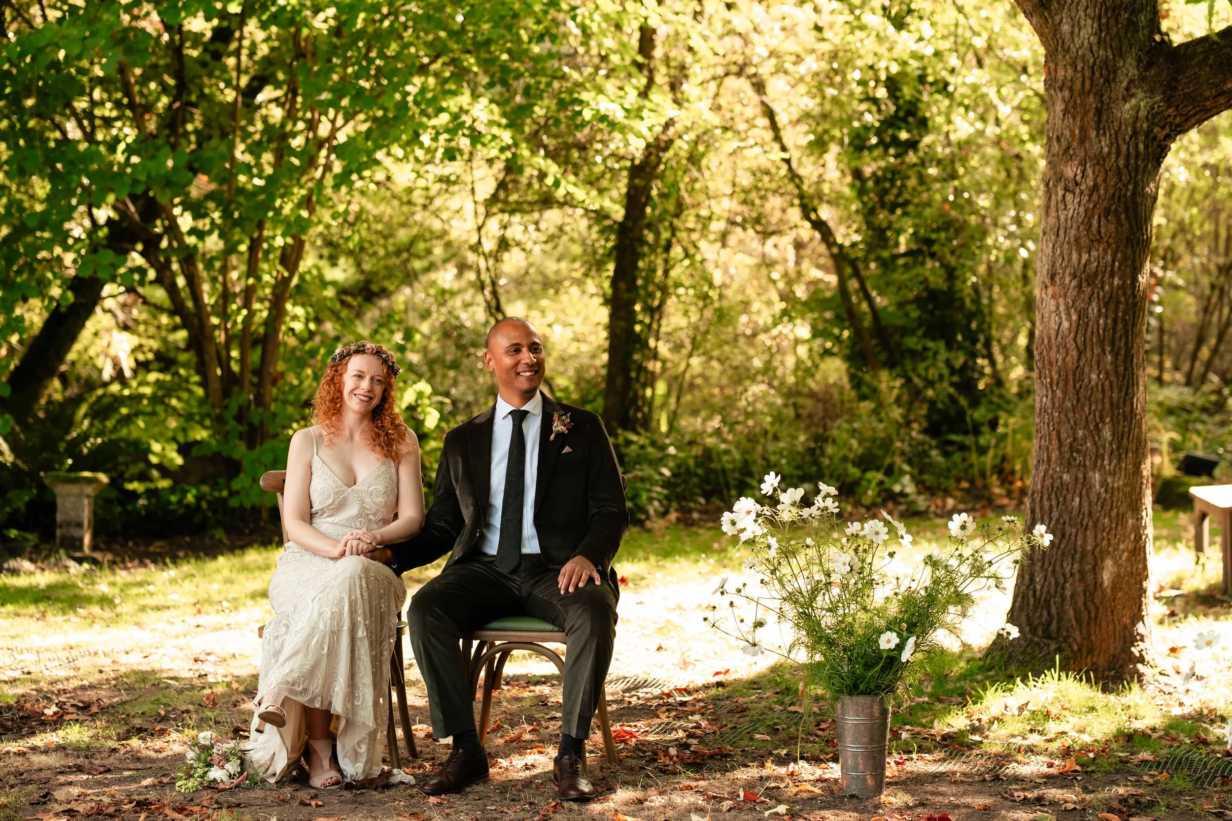 A bride and groom sitting outdoors on wedding chairs, holding hands, surrounded by greenery and trees with sunlight filtering through, with a vase of white flowers nearby.