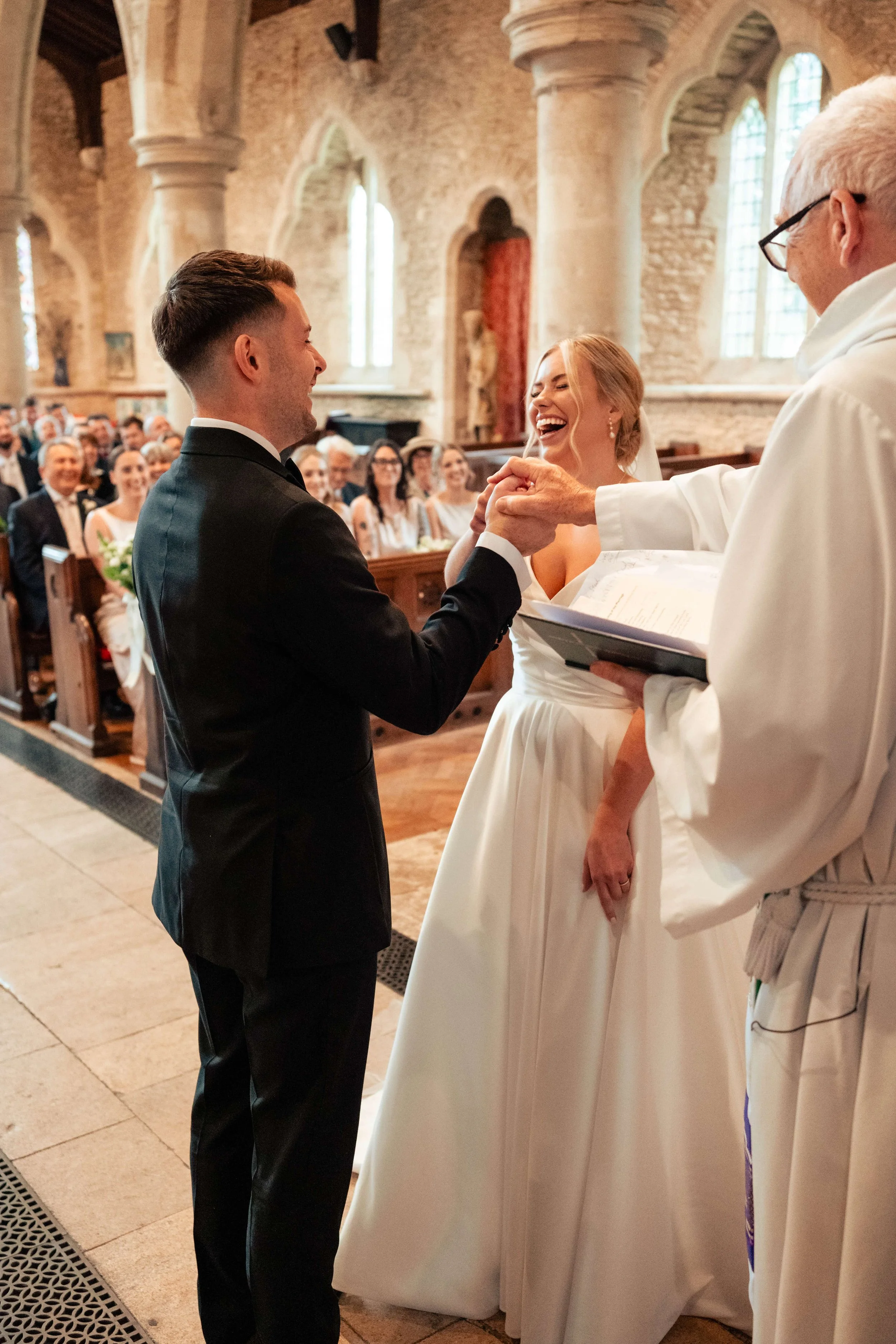 A wedding ceremony inside a church with a speaking priest, a bride and groom exchanging vows, and guests in the background.