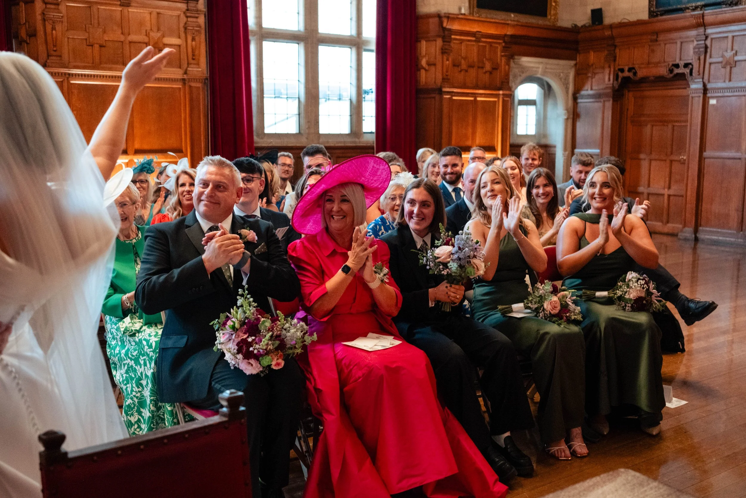 Wedding guests sitting in the Oxford Town Hall, clapping and smiling as they watch a person in a white dress gesturing, with some holding flowers, during a wedding ceremony.